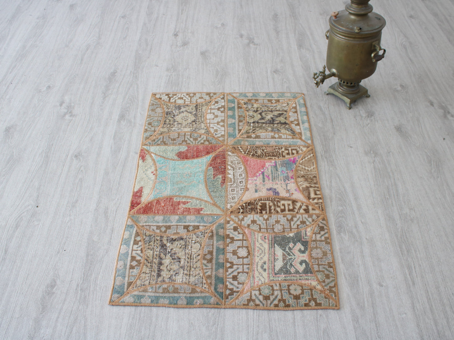 A rectangular vintage-style patchwork rug with a distressed geometric pattern in muted beige, brown, teal, and red tones, laid out on a light gray wood floor next to a brass samovar.