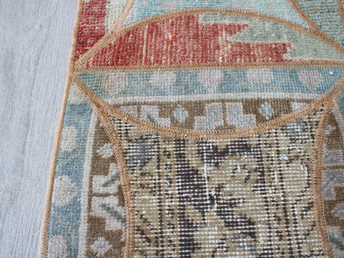 Close-up detail of a patterned area rug showing woven sections in beige, brown, light blue, and muted red, with a tan stitched border on a light wood floor.