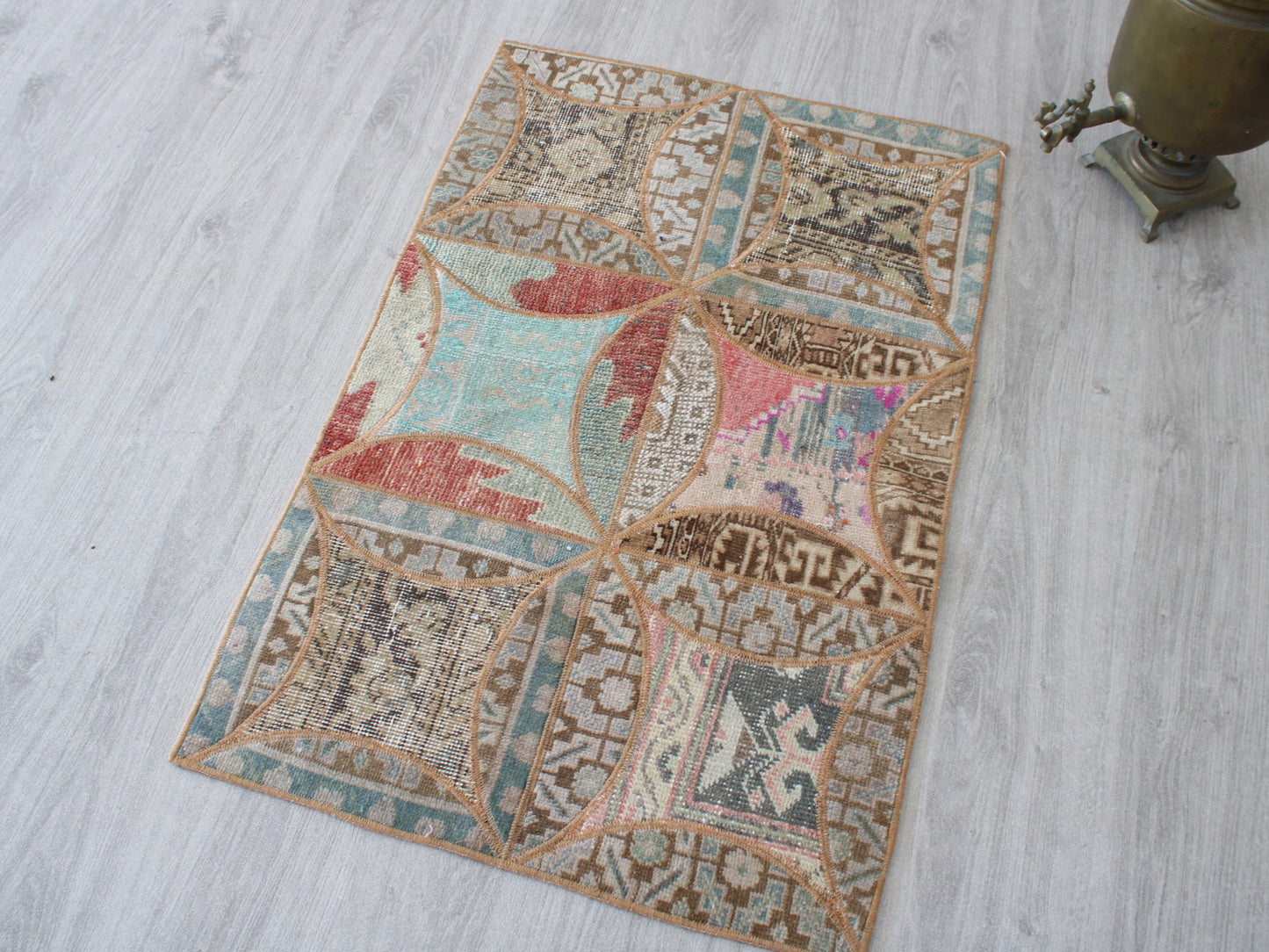 A rectangular vintage-style area rug with a patchwork design of geometric and floral motifs in muted teal, terracotta, pink, beige, and brown, laid out on a light gray wood floor next to a brass object.