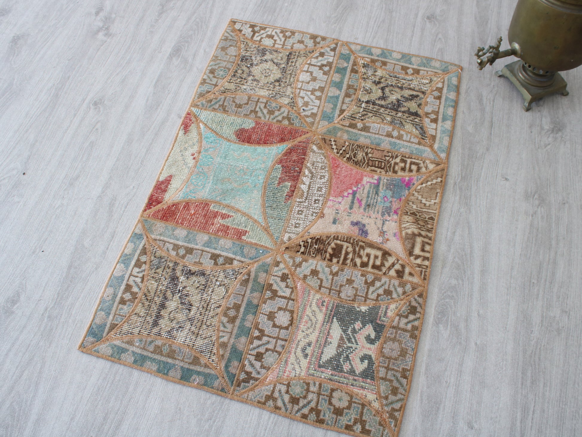 A rectangular vintage-style area rug with a patchwork design of geometric and floral motifs in muted teal, terracotta, pink, beige, and brown, laid out on a light gray wood floor next to a brass object.
