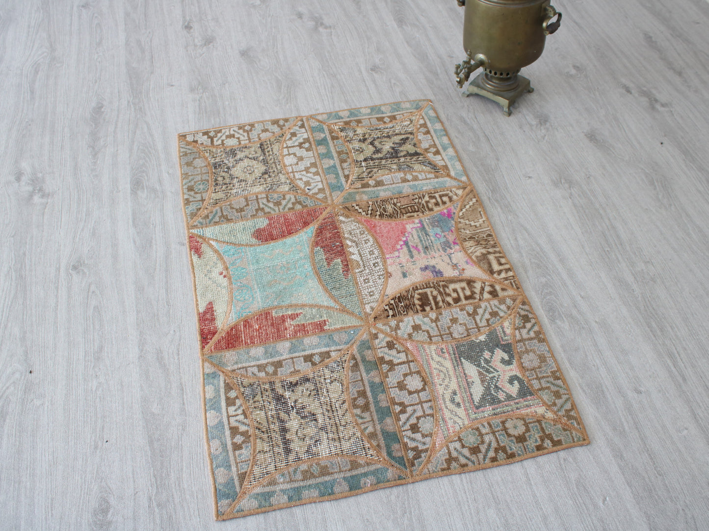 A rectangular vintage-style area rug with a patchwork of geometric and floral motifs in muted beige, teal, red, and brown tones, displayed on a light gray wood floor next to a brass samovar.