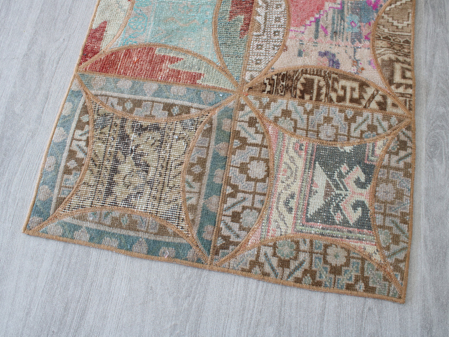 A close-up of a vintage-style patchwork area rug with geometric and floral motifs in muted shades of beige, brown, teal, pink, and red, laid out on a light gray wood floor.