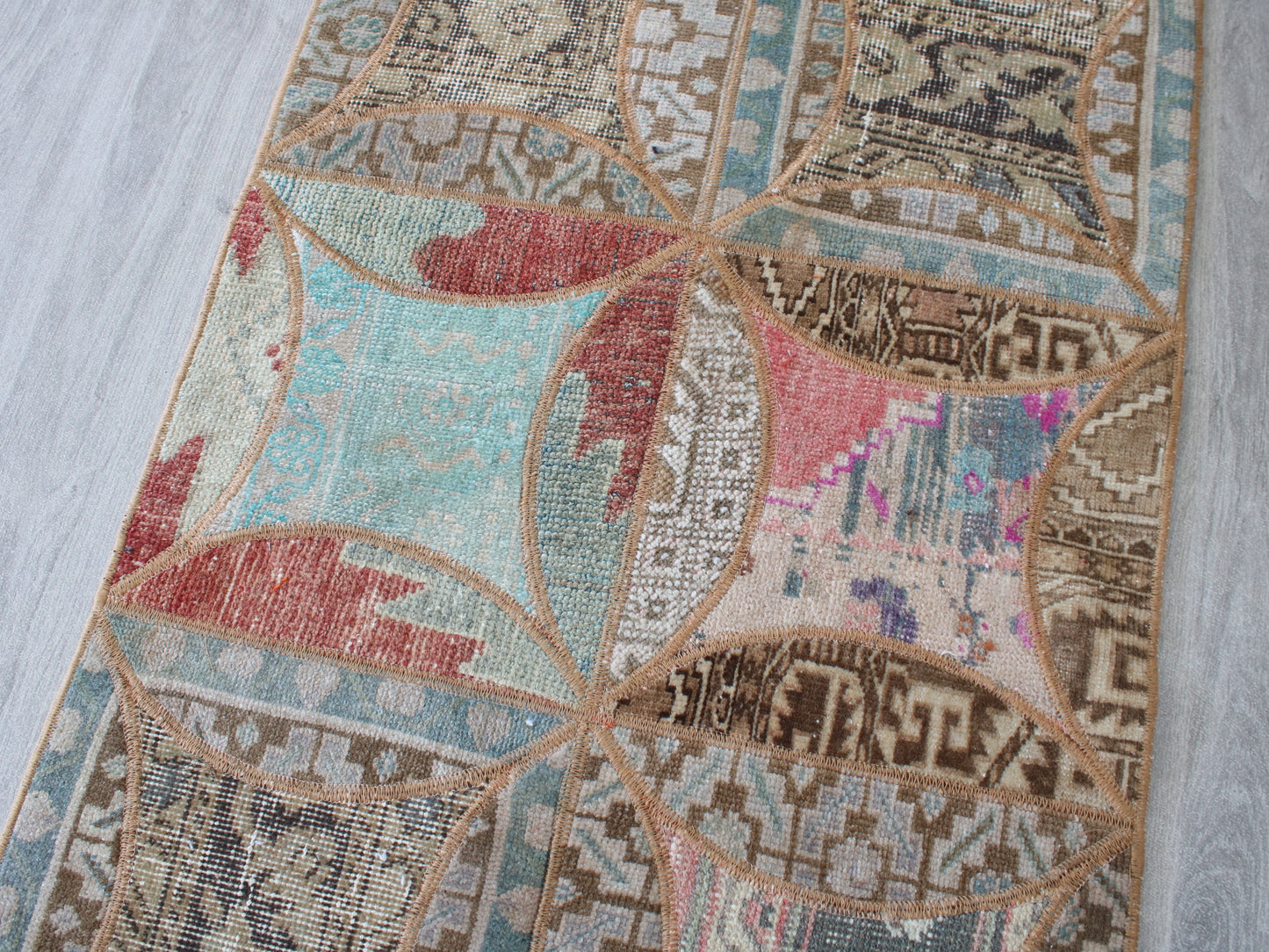 A close-up of a rectangular area rug laid on a light gray wood floor. The rug features a patchwork design with geometric and tribal-inspired patterns in beige, brown, light blue, muted red, and pink.