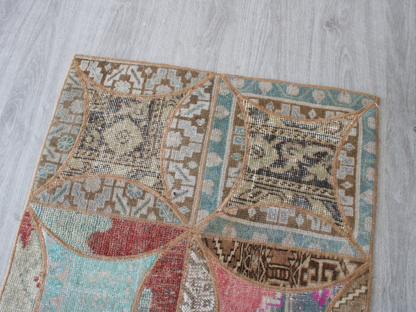 A square patchwork-style rug laid on a light gray wood floor, featuring multiple geometric and floral motifs in beige, brown, turquoise, red, pink, and cream.