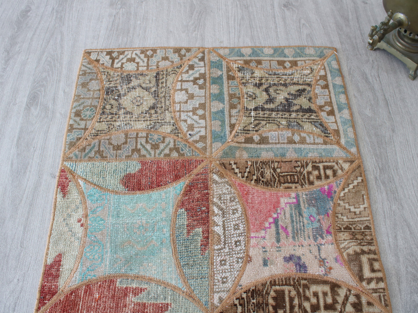 A square area rug with a distressed patchwork design, featuring geometric and tribal patterns in beige, brown, light blue, and muted red, laid out on a light gray wood floor.