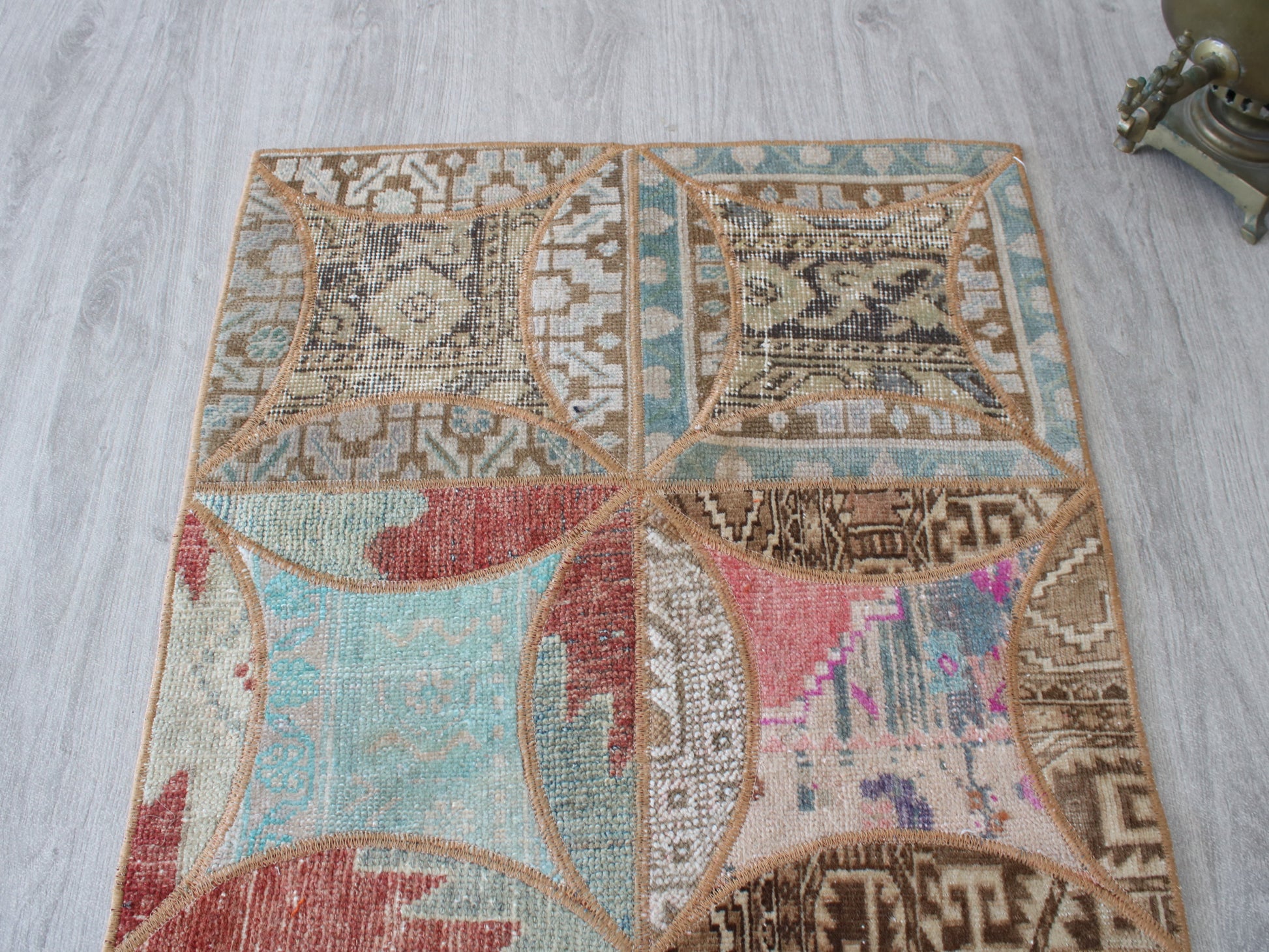 A square area rug with a distressed patchwork design, featuring geometric and tribal patterns in beige, brown, light blue, and muted red, laid out on a light gray wood floor.
