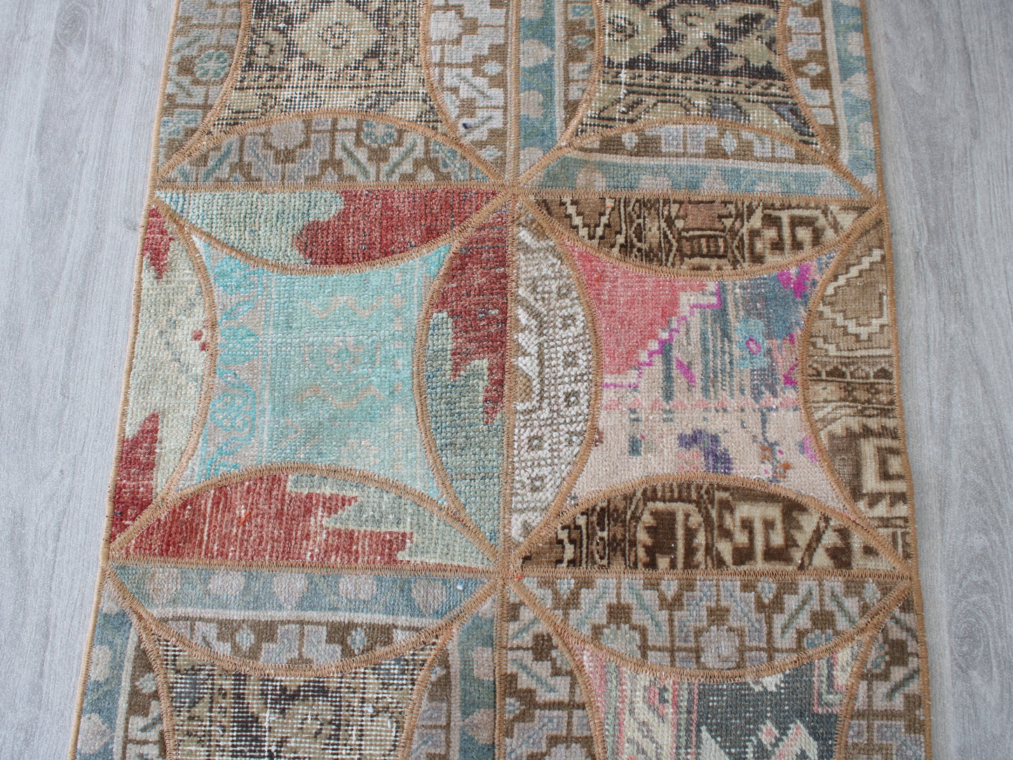 A close-up of a vintage-style geometric patchwork rug laid out on a light gray wood floor. The rug features a patchwork design with various tribal patterns in shades of beige, brown, light blue, dusty red, and pink, outlined in tan.