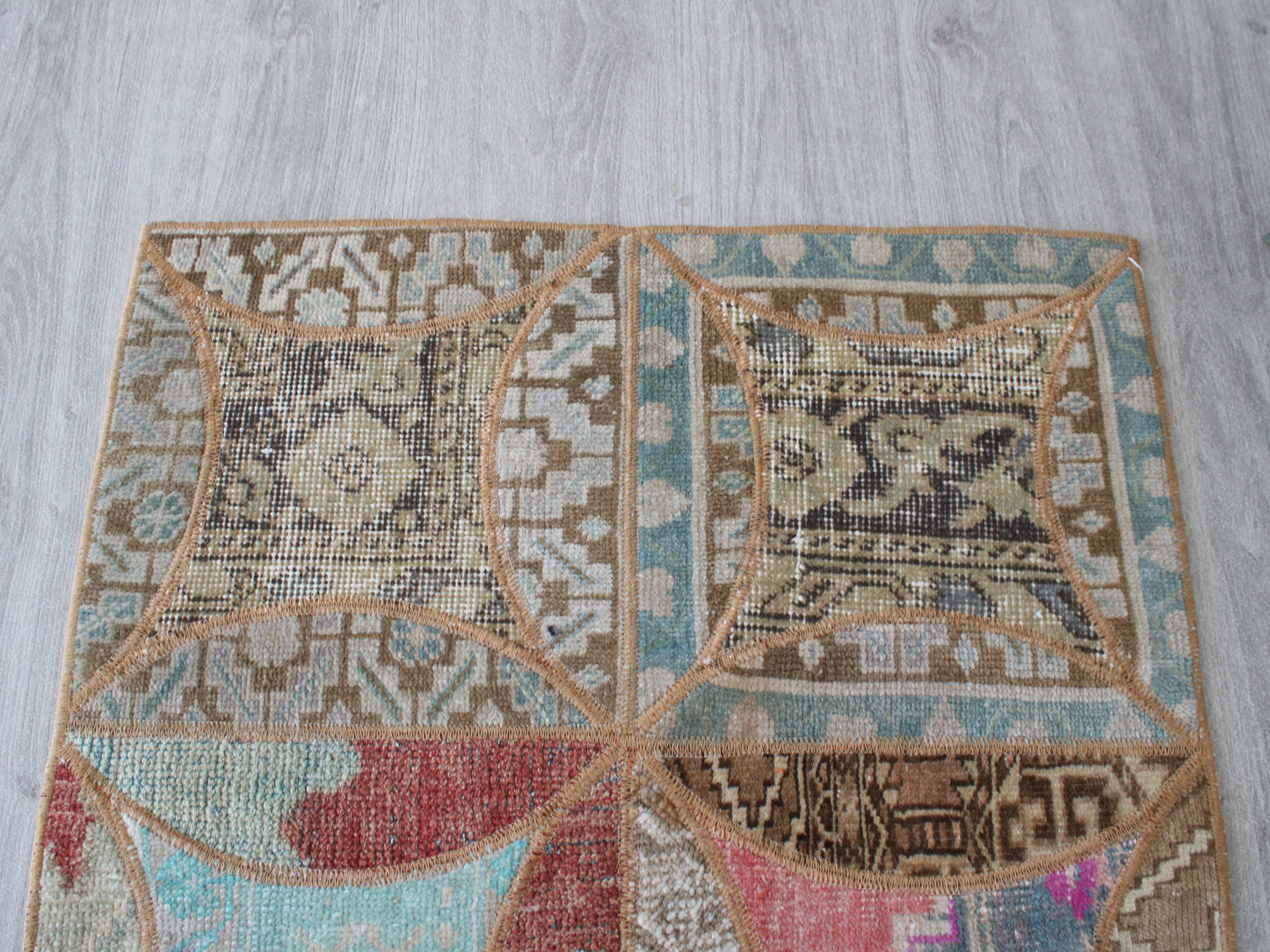A square area rug with a geometric patchwork design, laid flat on a light gray wood floor. The rug features a complex pattern of four large quadrants in beige, brown, light blue, and muted red, separated by tan stitched borders.