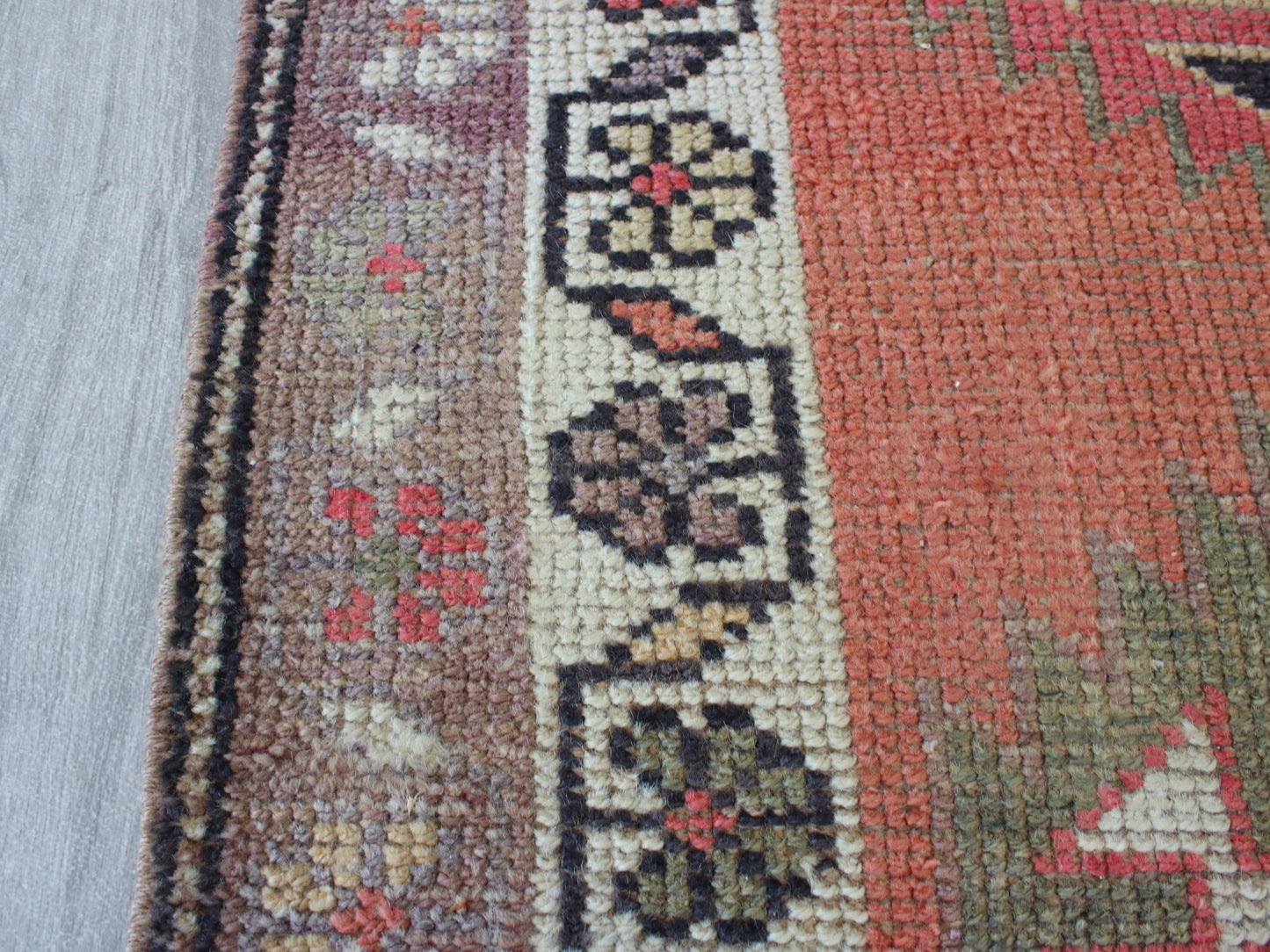 A close-up of a patterned area rug showing its woven texture, featuring a terracotta red field, a cream central band with floral motifs, and a brown side border.