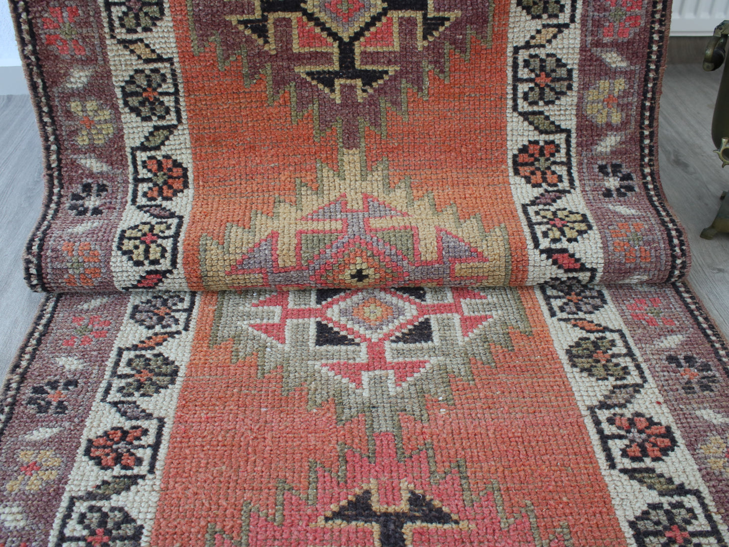 A vintage-style kilim runner rug with a distressed orange-red center, cream and pink geometric medallions, and a dark floral border, shown partially rolled up on a light-colored floor.