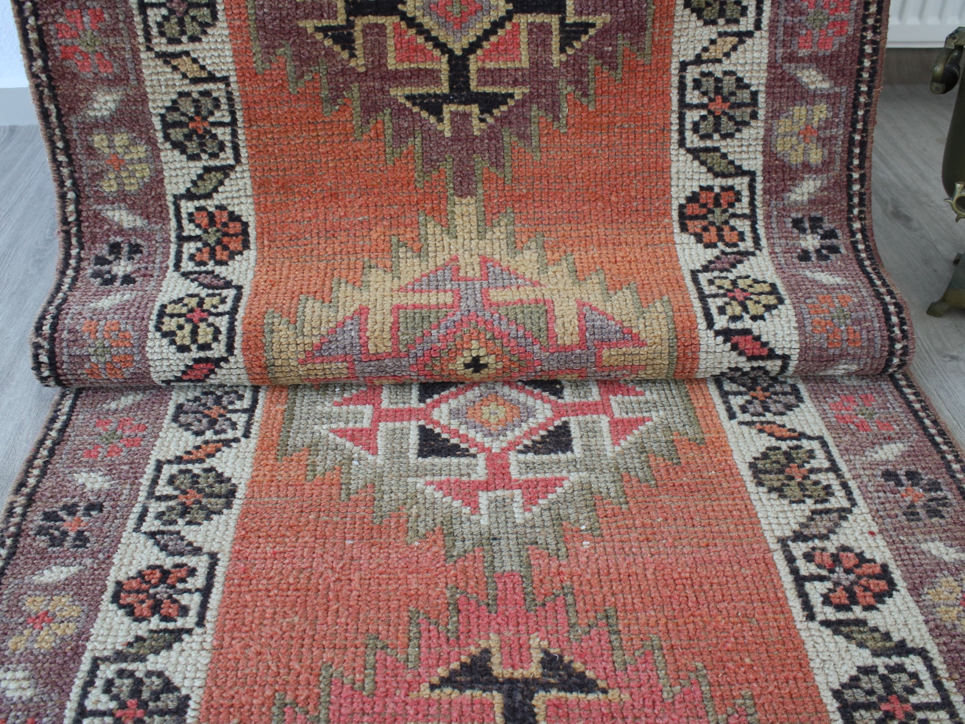 A vintage-style kilim runner rug with a distressed orange-red center, cream and pink geometric medallions, and a dark floral border, shown partially rolled up on a light-colored floor.
