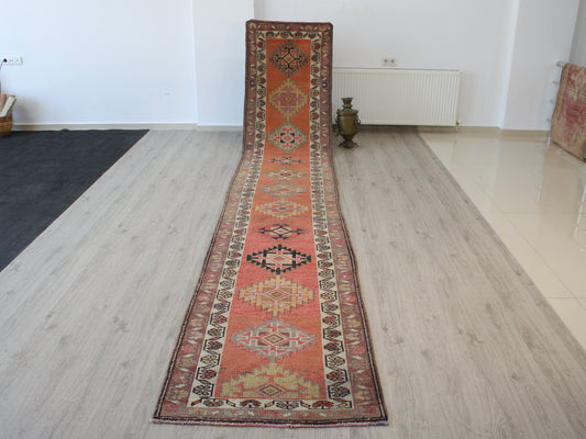 A long, narrow oriental runner rug with a terracotta orange-red base and intricate geometric patterns in beige, brown, and black, displayed on a light wood floor in a room.