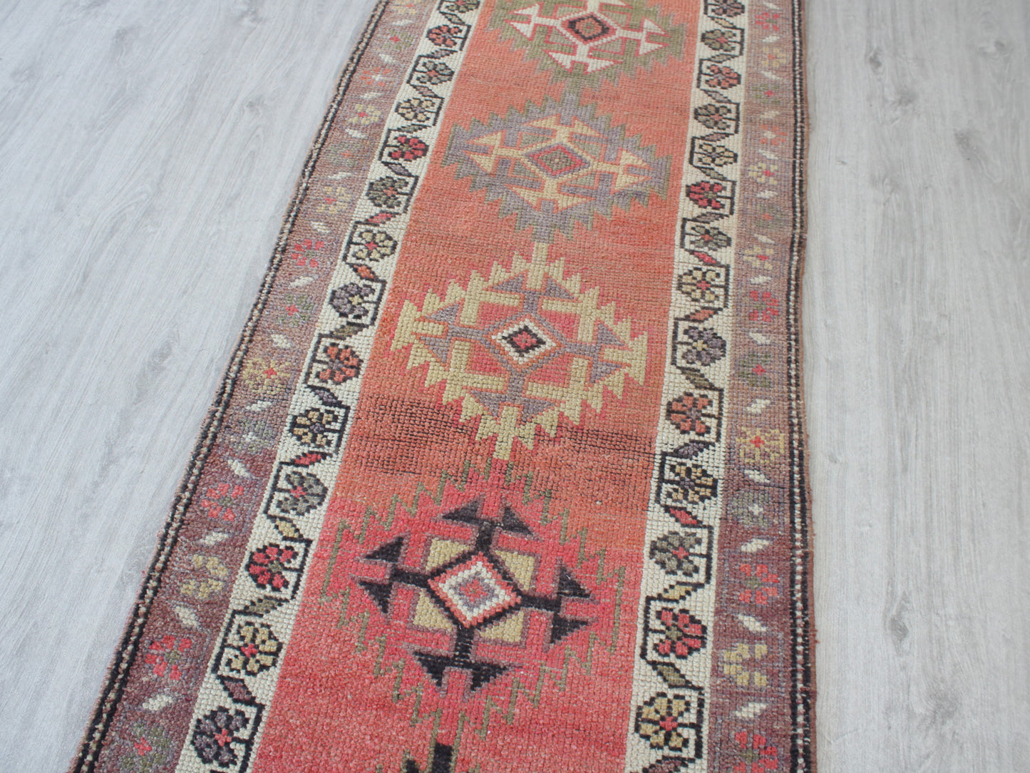 A long rectangular area rug with a distressed geometric pattern, featuring a terracotta red center, cream and beige diamond medallions, and dark brown borders, laid out on a light gray wood floor.