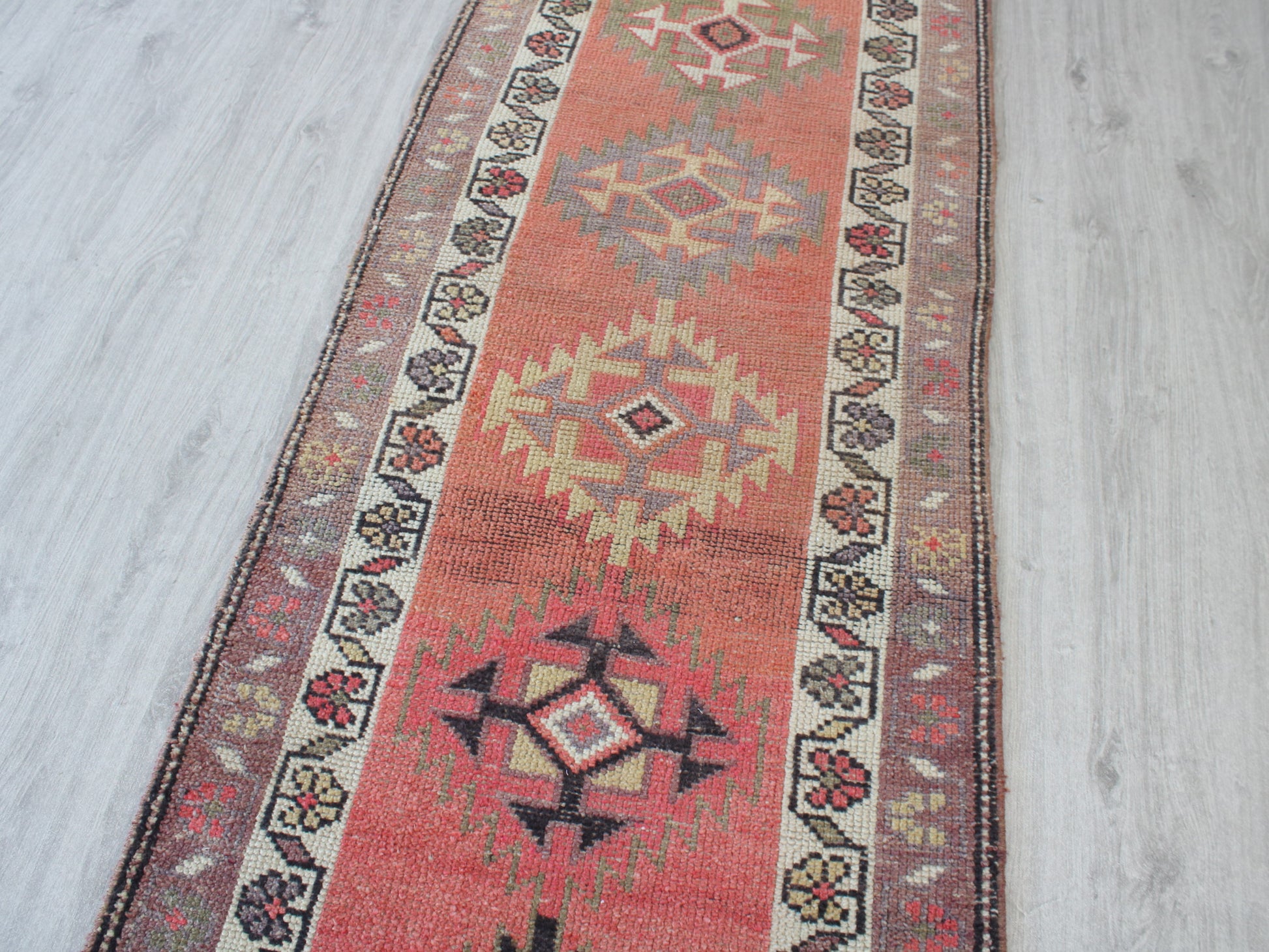 A long rectangular area rug with a distressed geometric pattern, featuring a terracotta red center, cream and beige diamond medallions, and dark brown borders, laid out on a light gray wood floor.