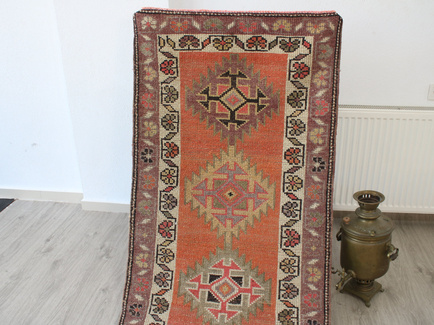 A traditional rectangular area rug with a warm orange-red center and geometric diamond motifs, leaning against a white wall on a light wood floor next to a brass samovar.