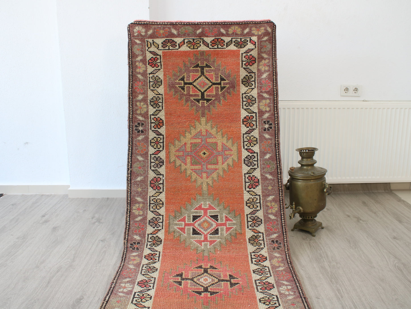 A long rectangular area rug with a traditional geometric pattern in terracotta orange, beige, and dark brown, displayed on a light wood floor next to a brass samovar in a minimalist room.