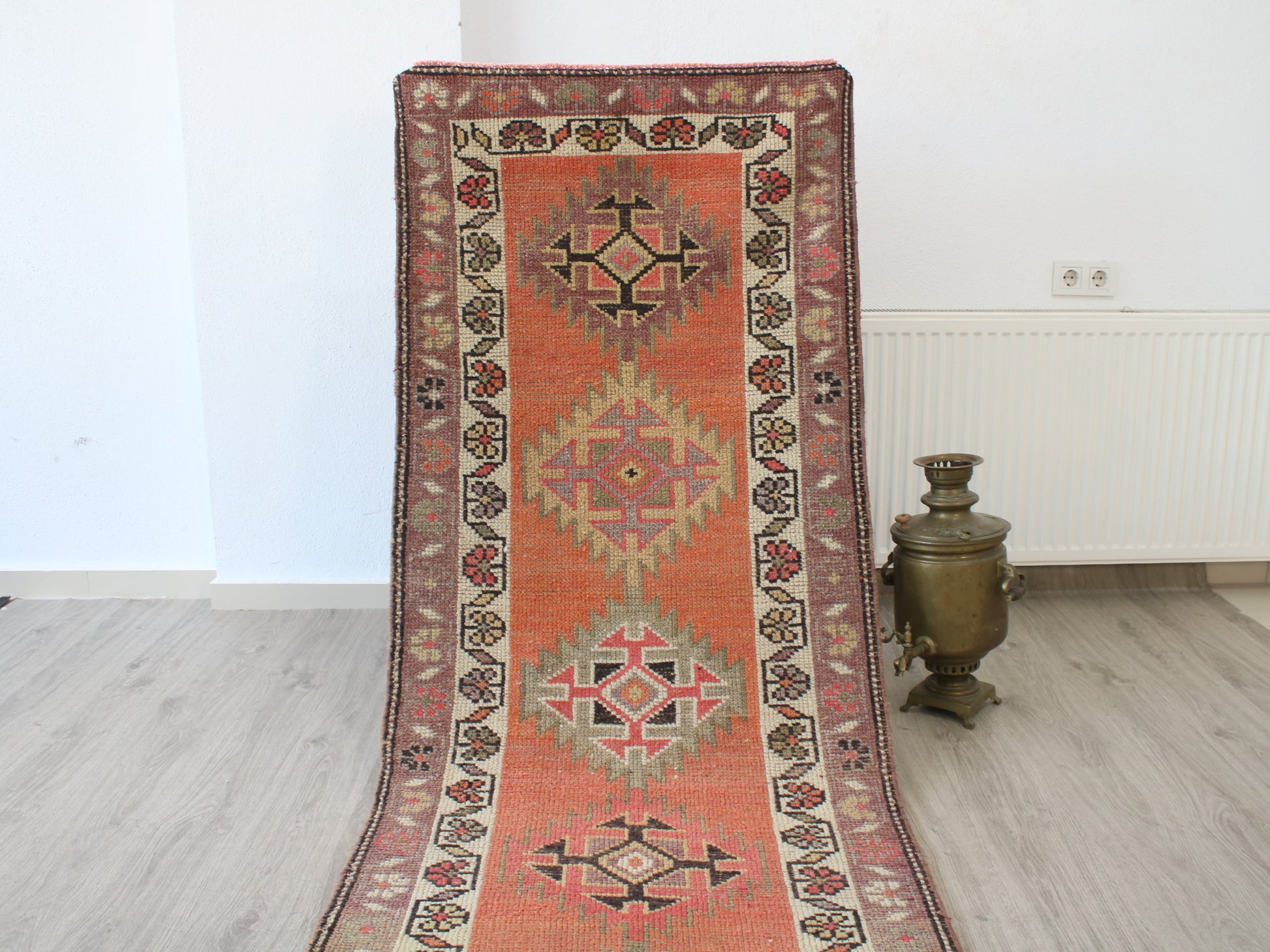 A long rectangular area rug with a traditional geometric pattern in terracotta orange, beige, and dark brown, displayed on a light wood floor next to a brass samovar in a minimalist room.