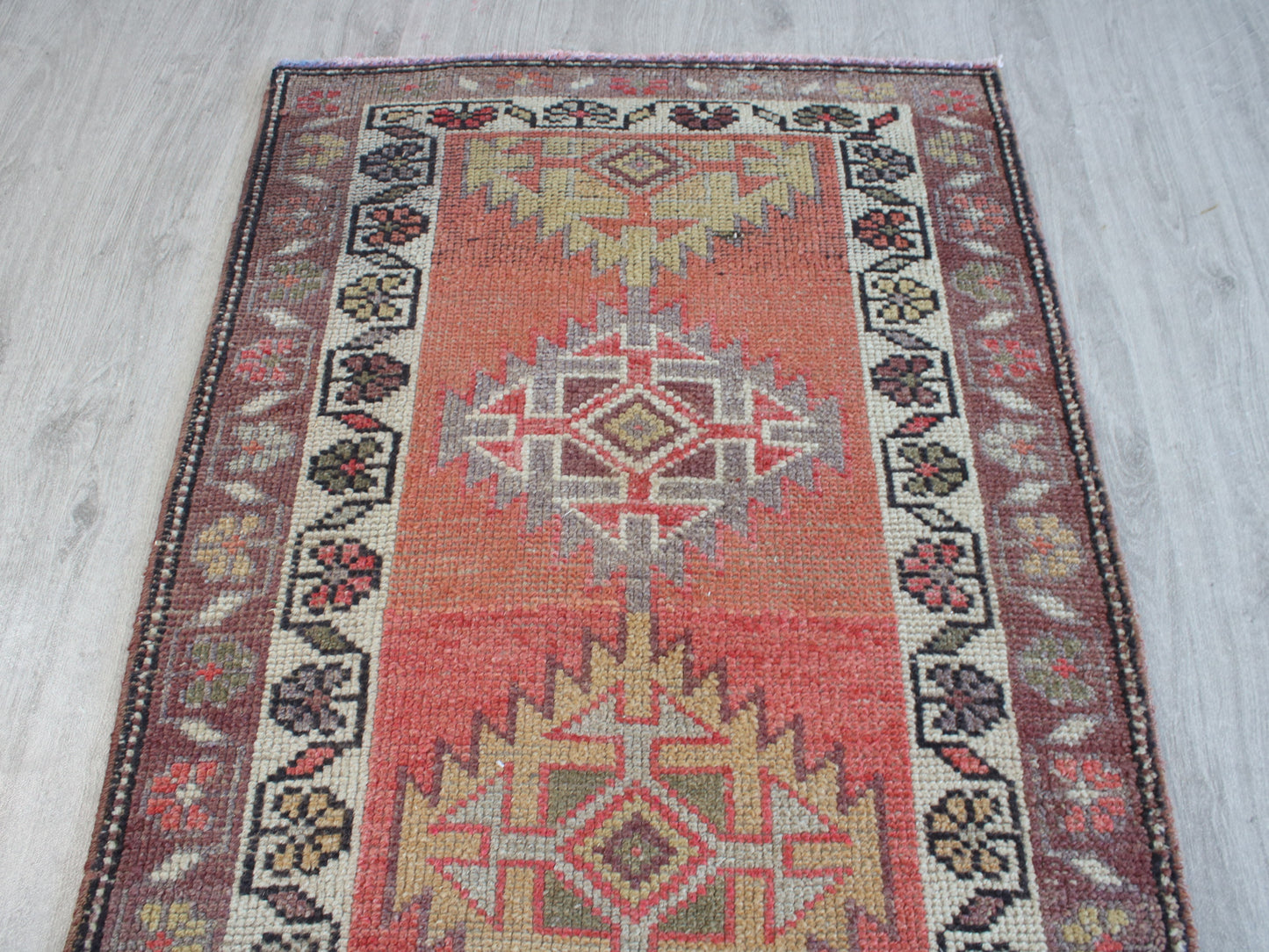 A rectangular vintage-style area rug with a distressed geometric pattern, featuring a central red field, beige and gray medallions, and a dark patterned border, laid flat on a light gray wood floor.