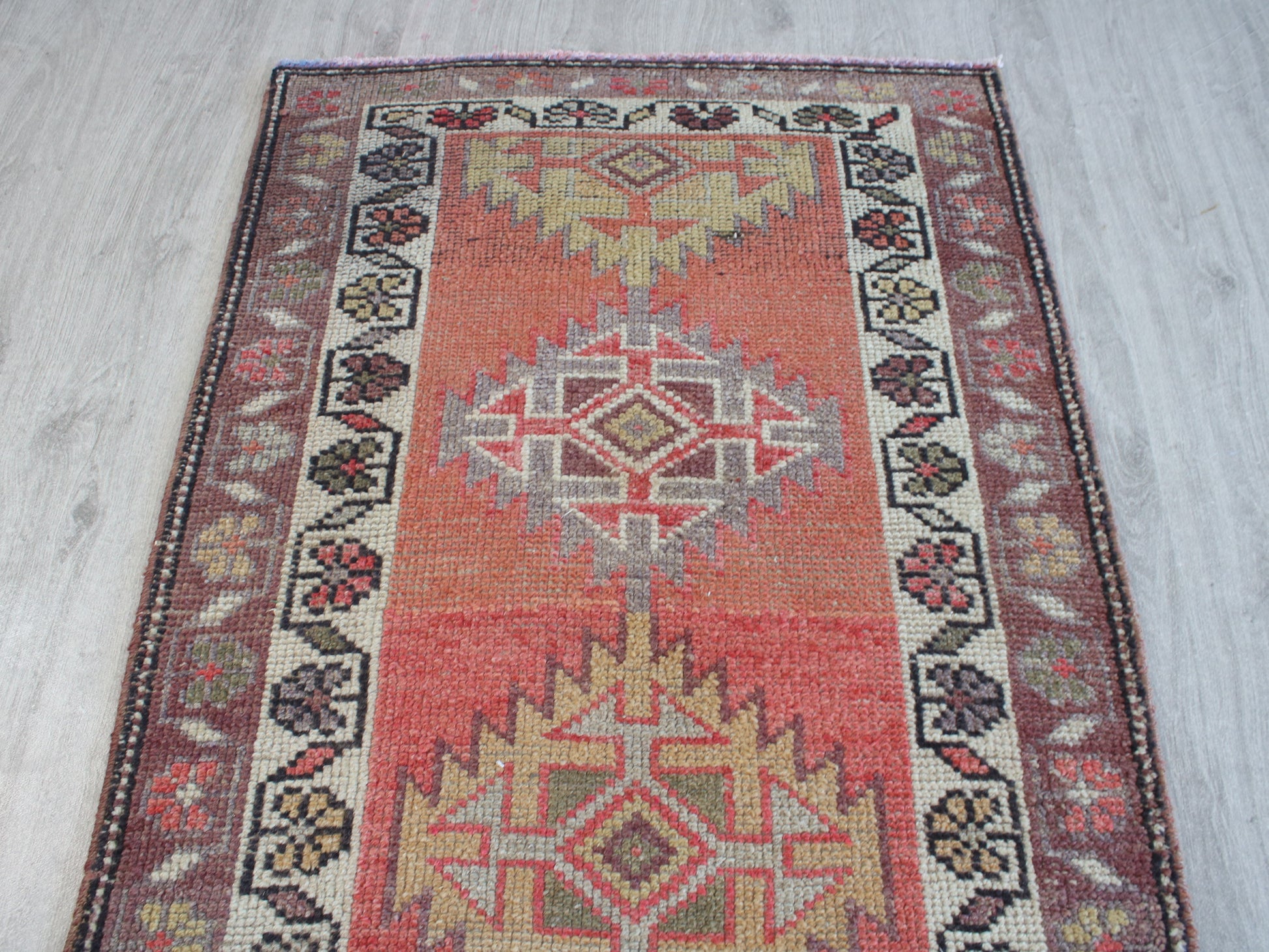 A rectangular vintage-style area rug with a distressed geometric pattern, featuring a central red field, beige and gray medallions, and a dark patterned border, laid flat on a light gray wood floor.