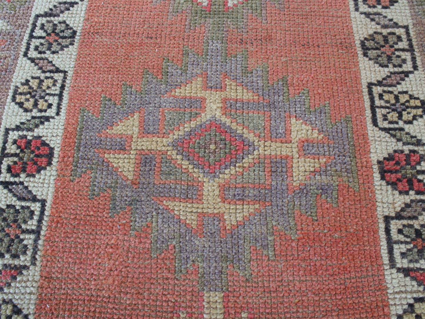 A close-up of a woven rug with a central geometric diamond motif in muted coral, beige, gray, and red, bordered by floral patterns on cream and dark navy.