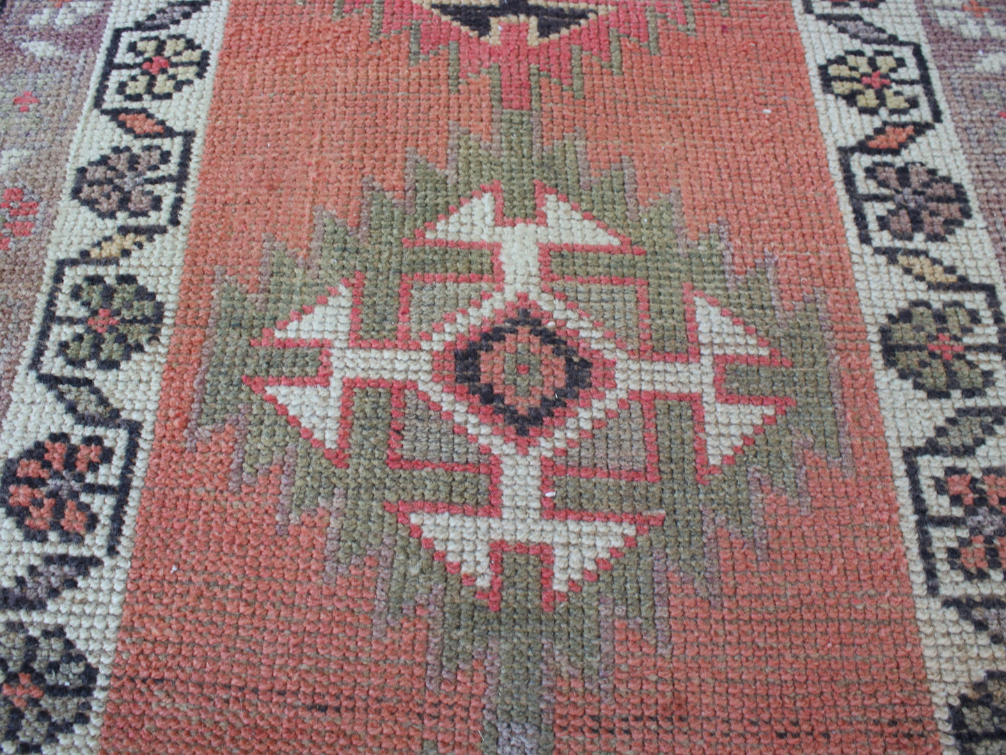 A close-up of a woven rug with a central geometric medallion pattern in muted coral, sage green, cream, and dark charcoal tones.