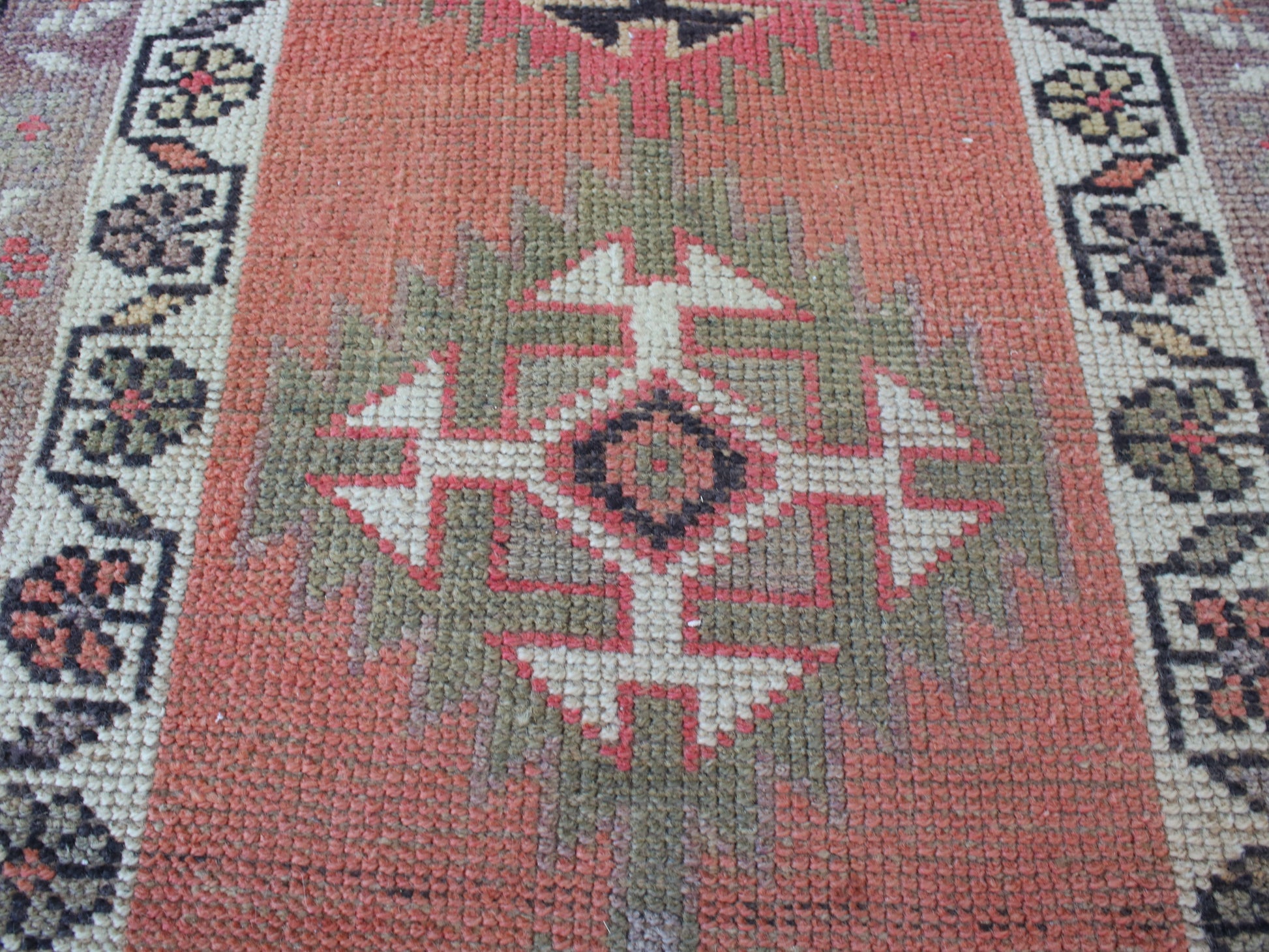 A close-up of a woven rug with a central geometric medallion pattern in muted coral, sage green, cream, and dark charcoal tones.