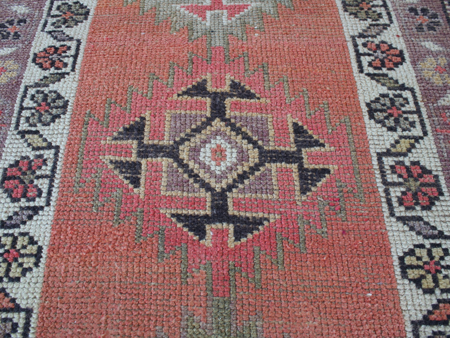 A close-up of a woven geometric rug with a terracotta red center field, black and beige central medallion, and patterned borders in muted green, cream, and burgundy.