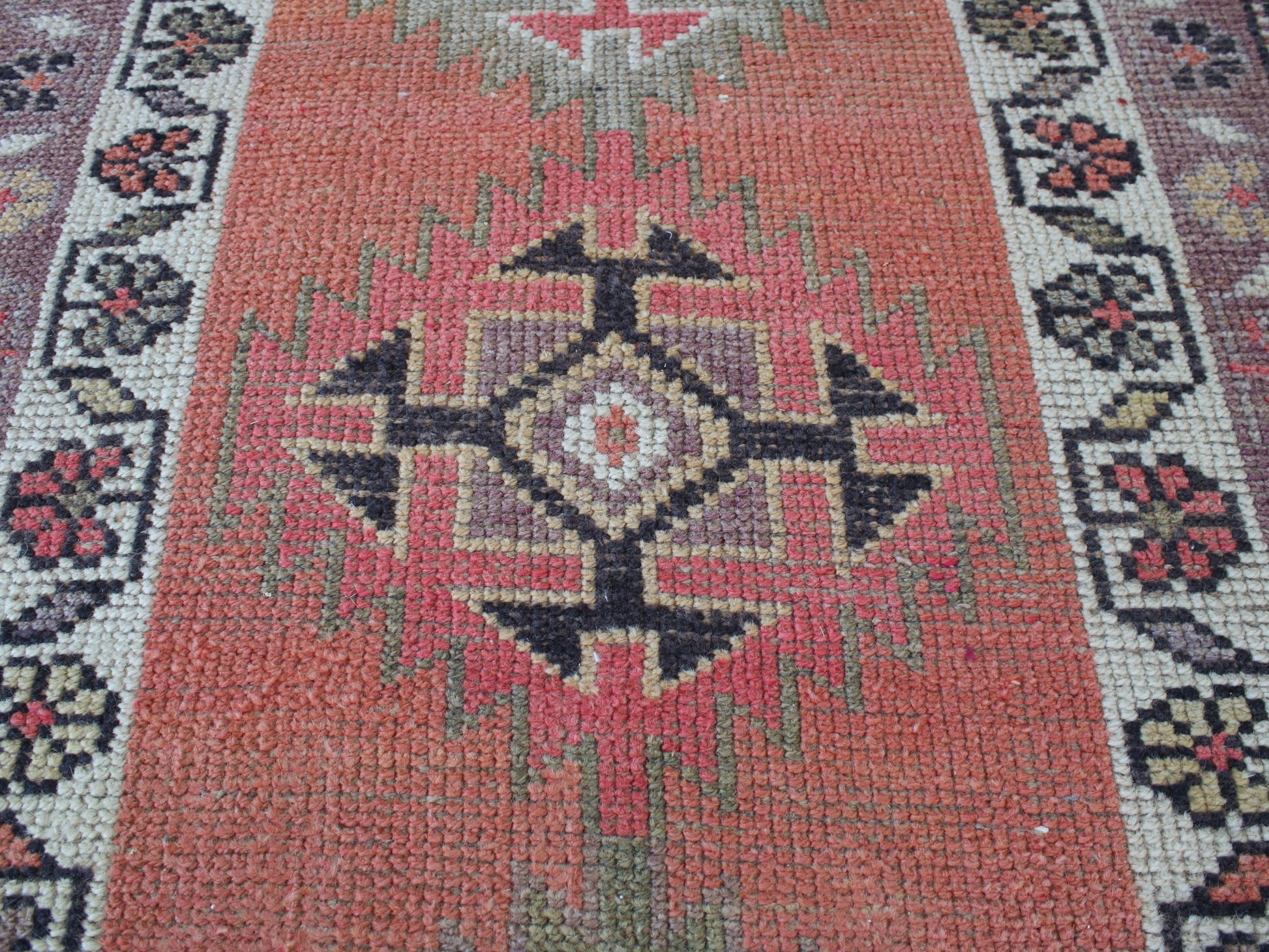 A close-up of a woven geometric rug with a terracotta red center field, black and beige central medallion, and patterned borders in muted green, cream, and burgundy.