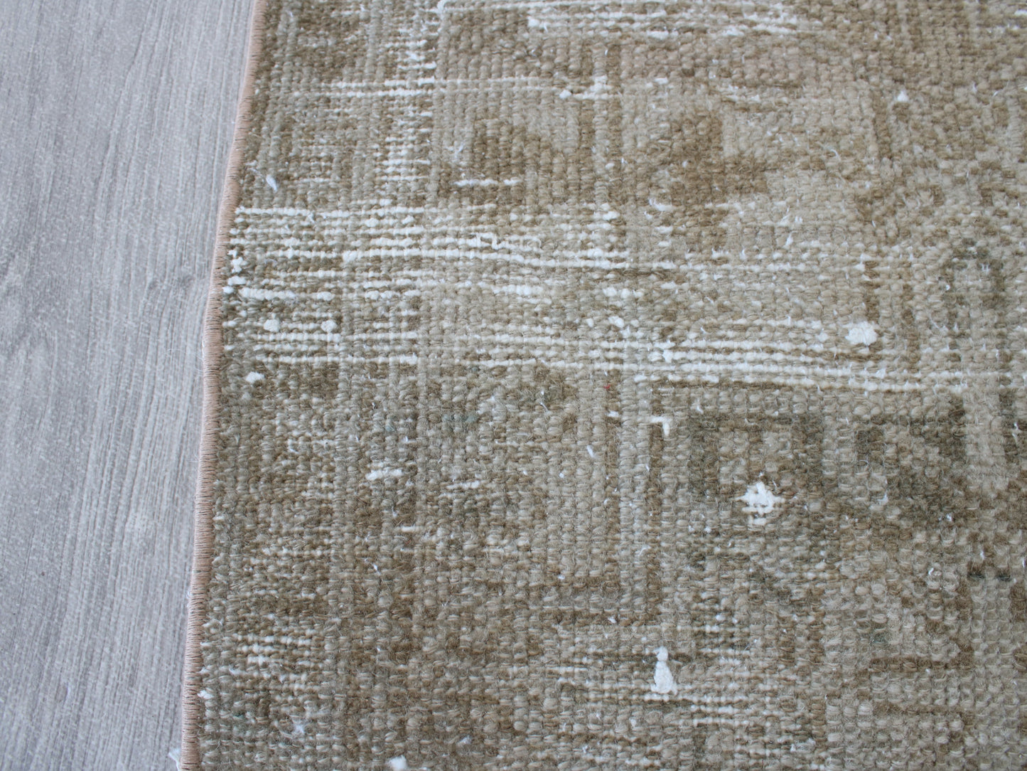 A close-up of a distressed area rug with a beige and brown geometric pattern, shown next to a light gray wood floor.