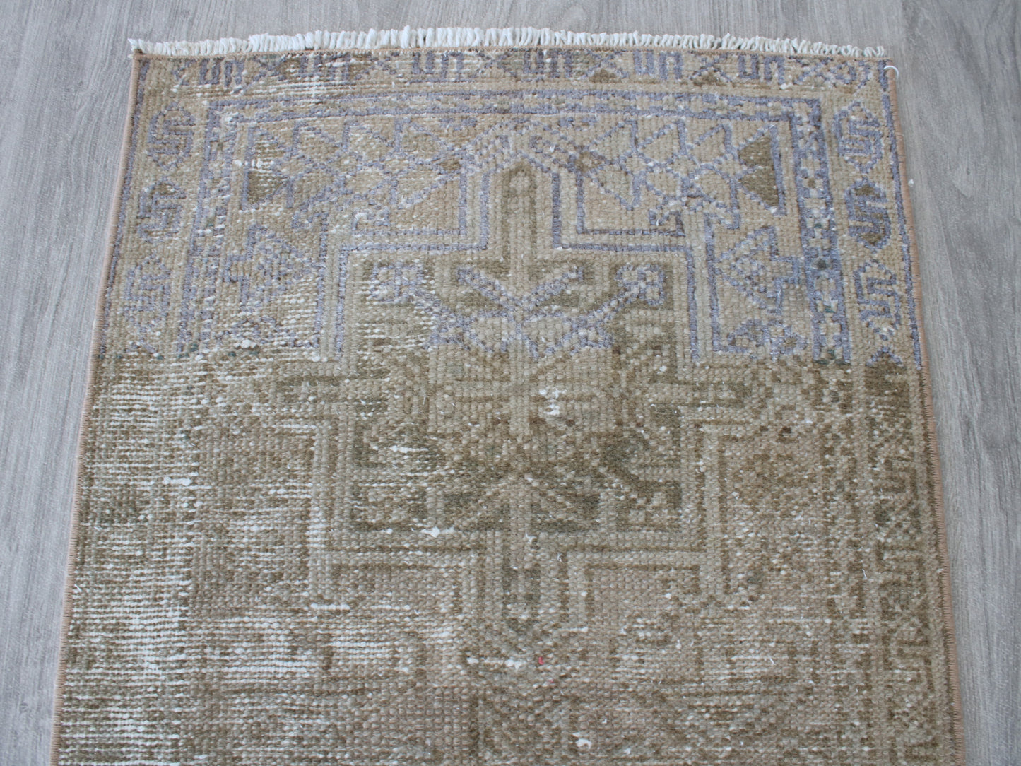 A square distressed area rug with a faded geometric medallion pattern in beige, brown, and muted blue-gray tones, featuring white fringe on the top edge, laid flat on a gray wood floor.