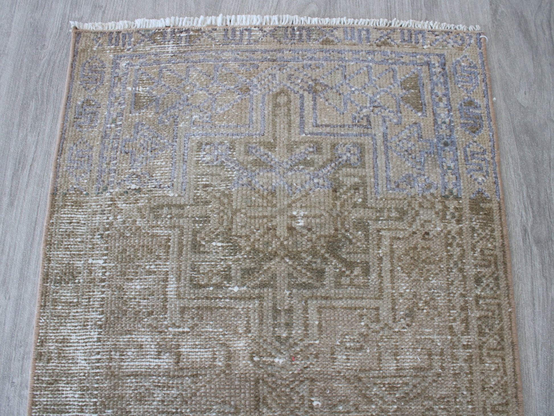 A square distressed area rug with a faded geometric medallion pattern in beige, brown, and muted blue-gray tones, featuring white fringe on the top edge, laid flat on a gray wood floor.