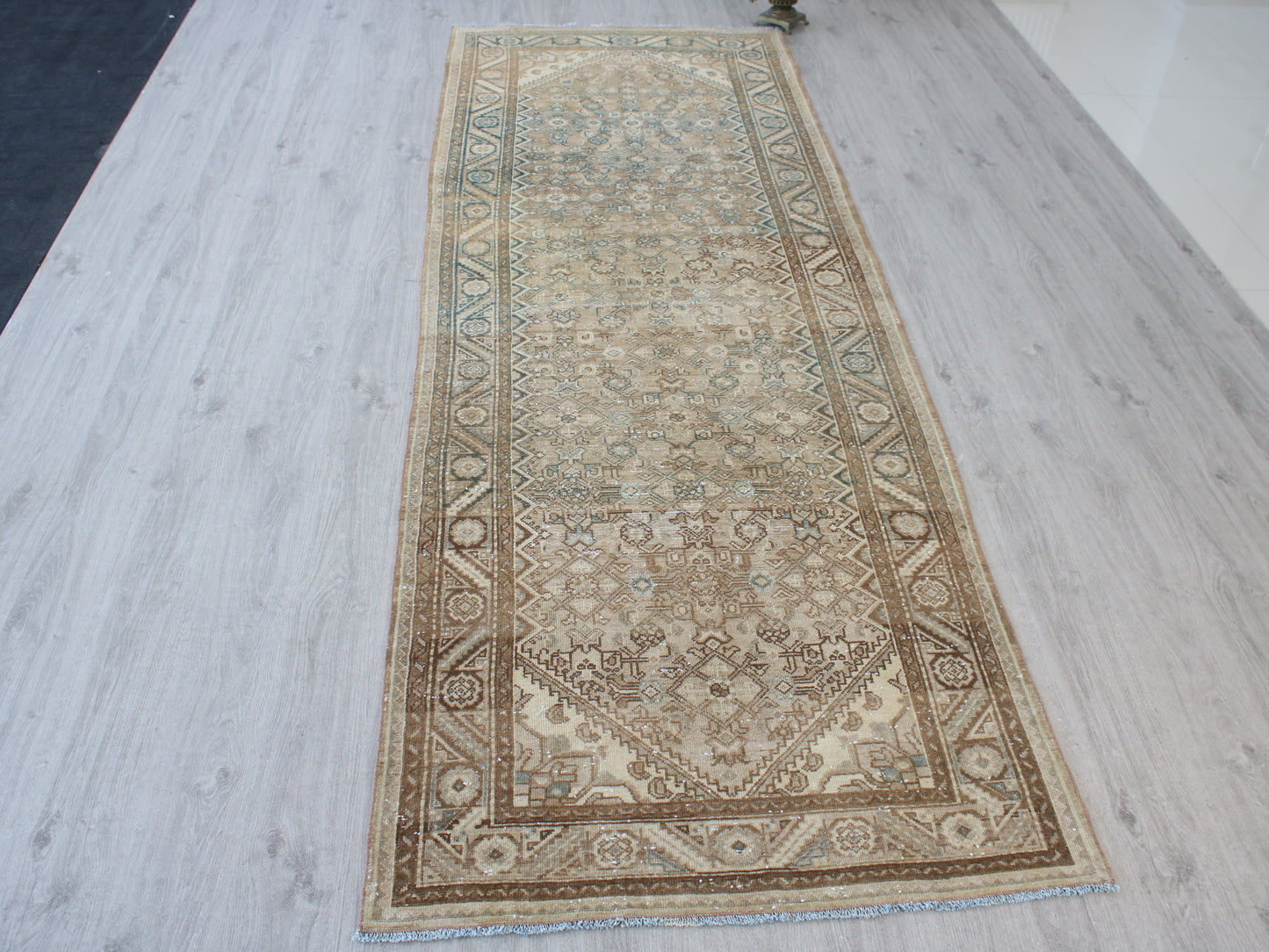 A long rectangular area rug with an intricate traditional pattern in beige, brown, and muted blue, laid out on a light gray wood-plank floor.