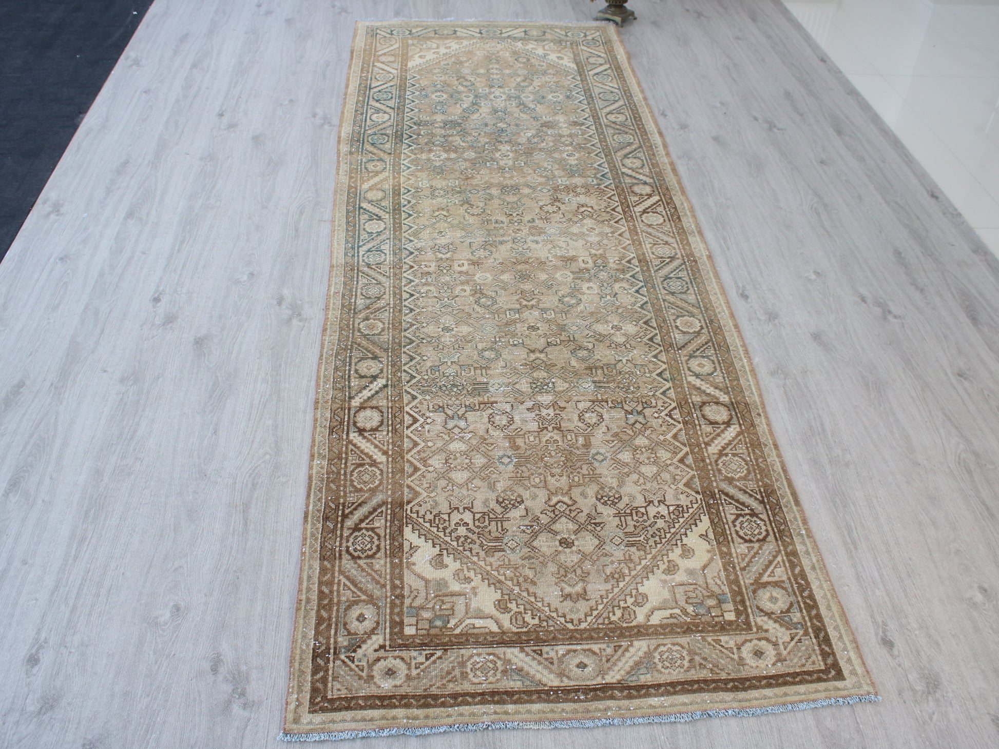 A long rectangular area rug with an intricate traditional pattern in beige, brown, and muted blue, laid out on a light gray wood-plank floor.
