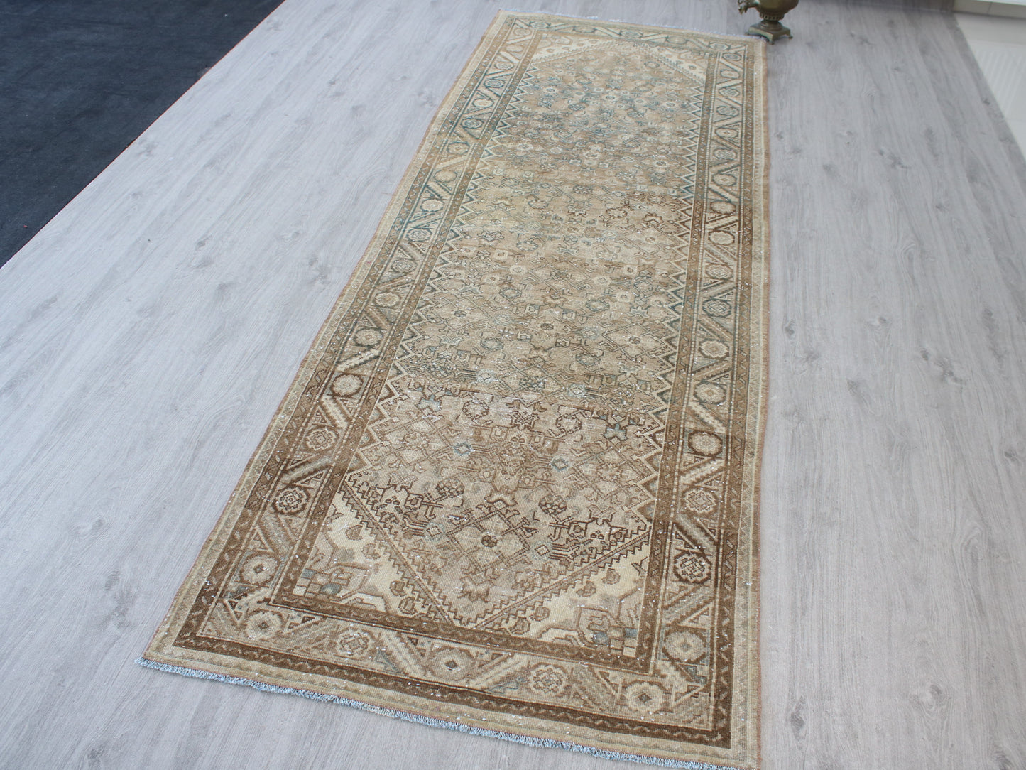 A long rectangular area rug with an intricate traditional floral pattern in beige, brown, and muted blue-green tones, laid out on a light gray wood-plank floor.