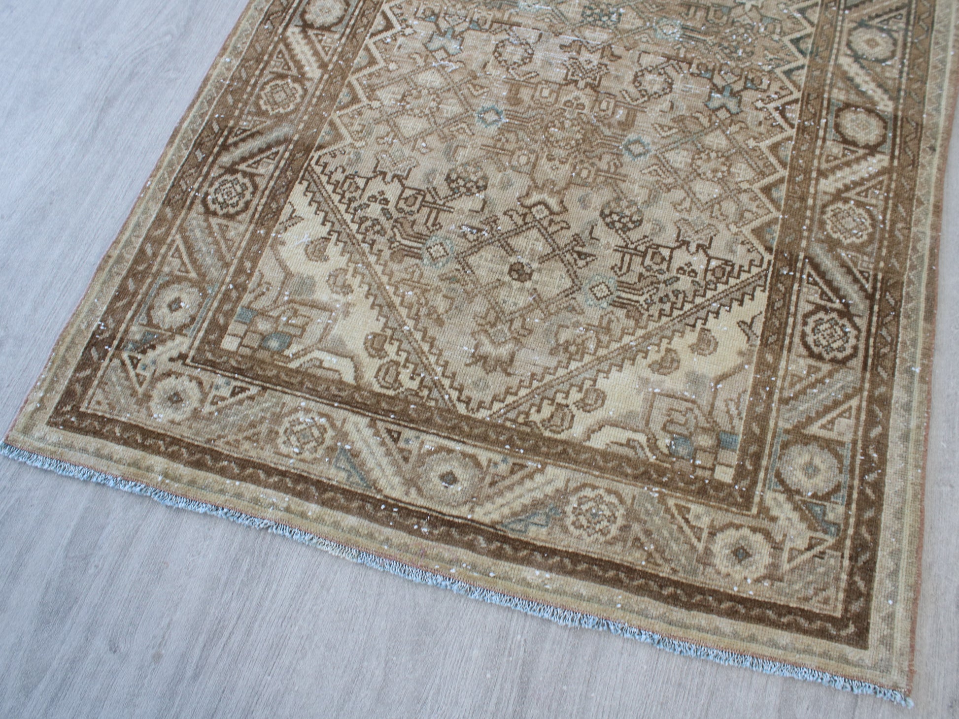 A traditional oriental-style area rug with an intricate geometric and floral pattern, shown laid out on a light gray wood floor. The rug features a warm beige and brown color palette with subtle light blue accents.