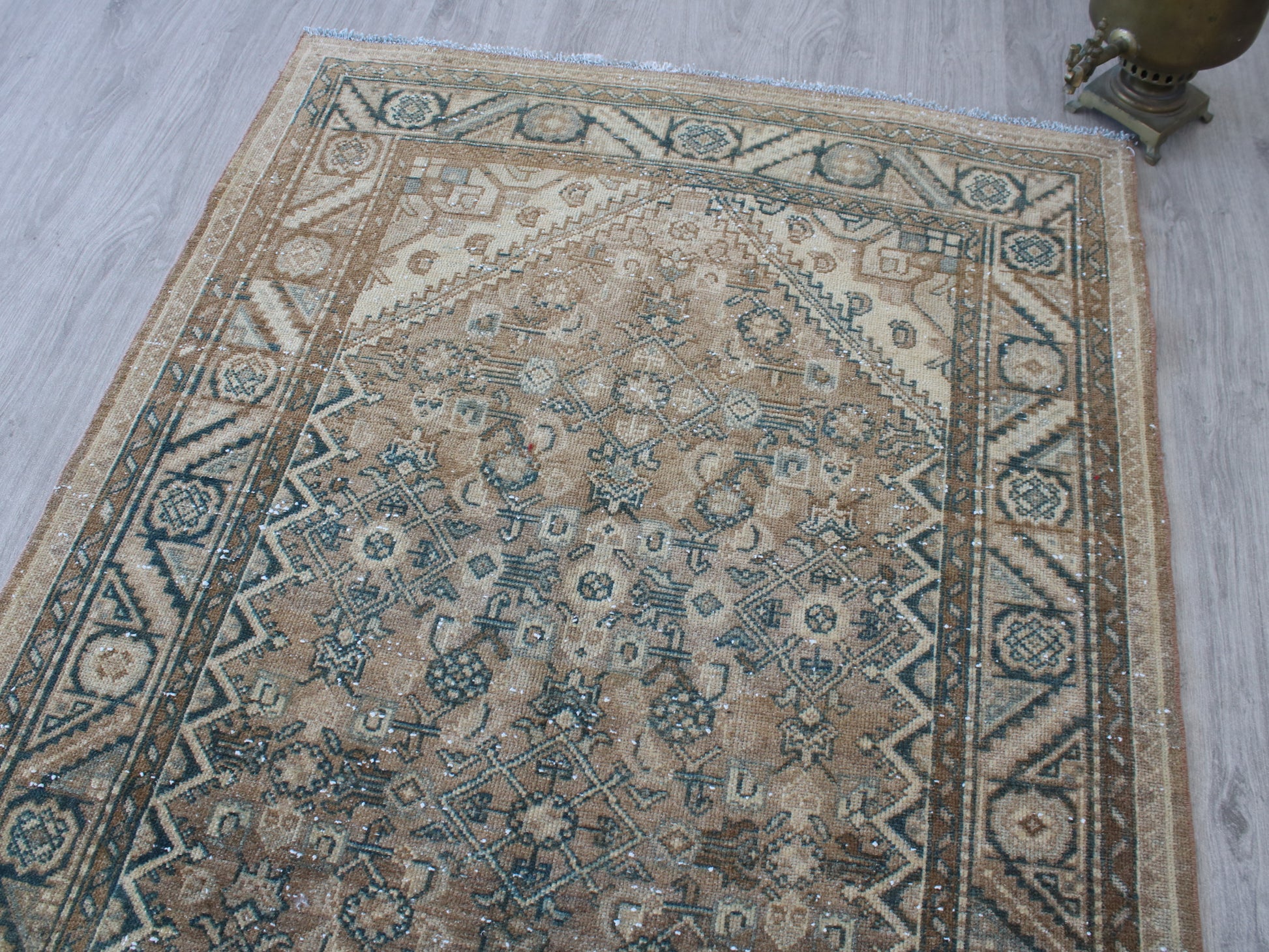 A rectangular vintage-style area rug with an intricate traditional floral and geometric pattern, shown laid out on a light gray wood floor. The rug features a warm beige base with dusty blue, brown, and cream patterns.