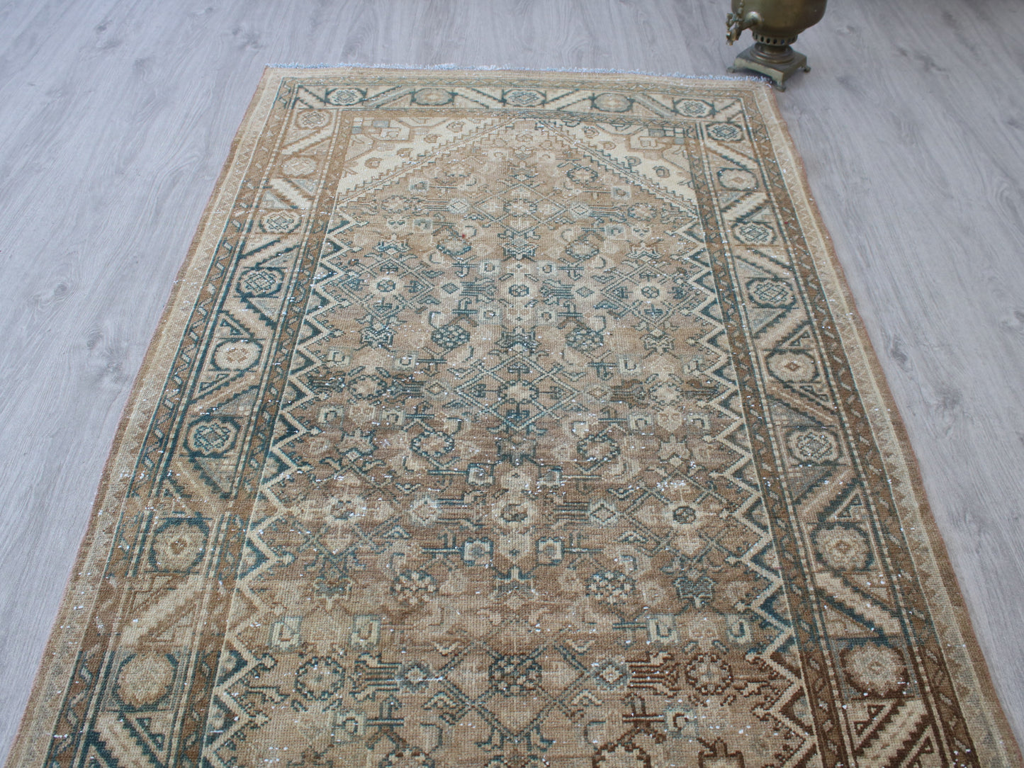 A rectangular vintage-style area rug with a distressed traditional pattern in beige, brown, and muted blue-green, laid out on a light gray wood floor.