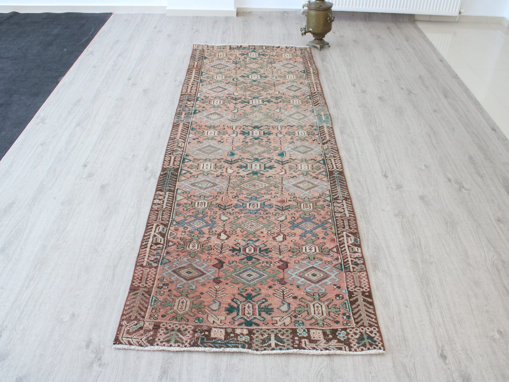 A long rectangular area rug with an intricate traditional geometric pattern in dusty pink, beige, brown, and teal, laid out on a light gray wood floor.