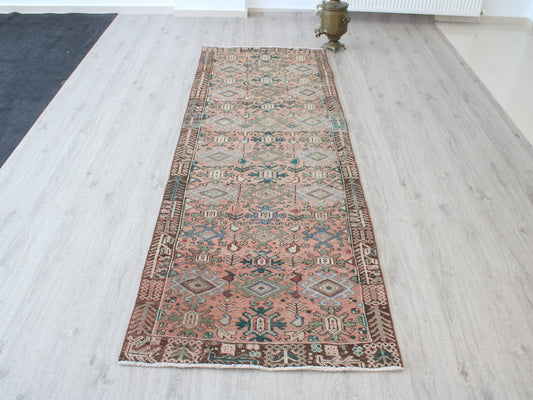 A long rectangular area rug with an intricate traditional geometric pattern in dusty pink, beige, brown, and teal, laid out on a light gray wood floor.