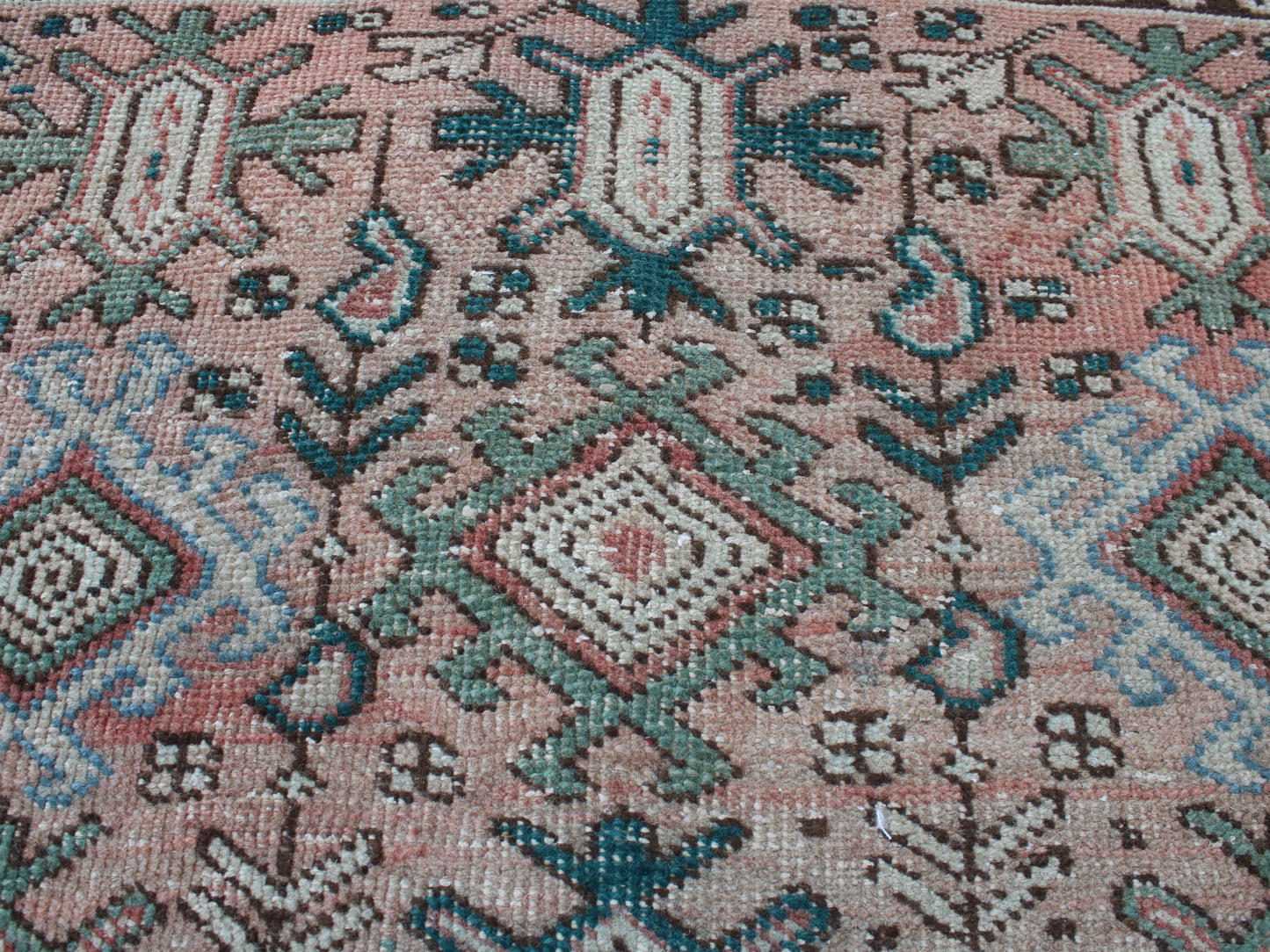 A close-up of a woven rug or textile with a geometric pattern in dusty pink, teal blue, sage green, and cream.