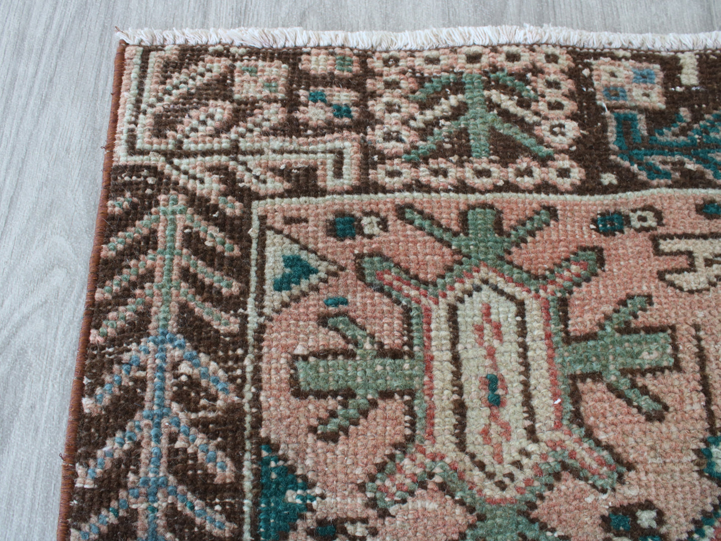 A close-up of a patterned area rug corner showing a dusty pink field with green, brown, and cream geometric motifs, with white fringe visible at the top edge, placed on a light gray wood floor.