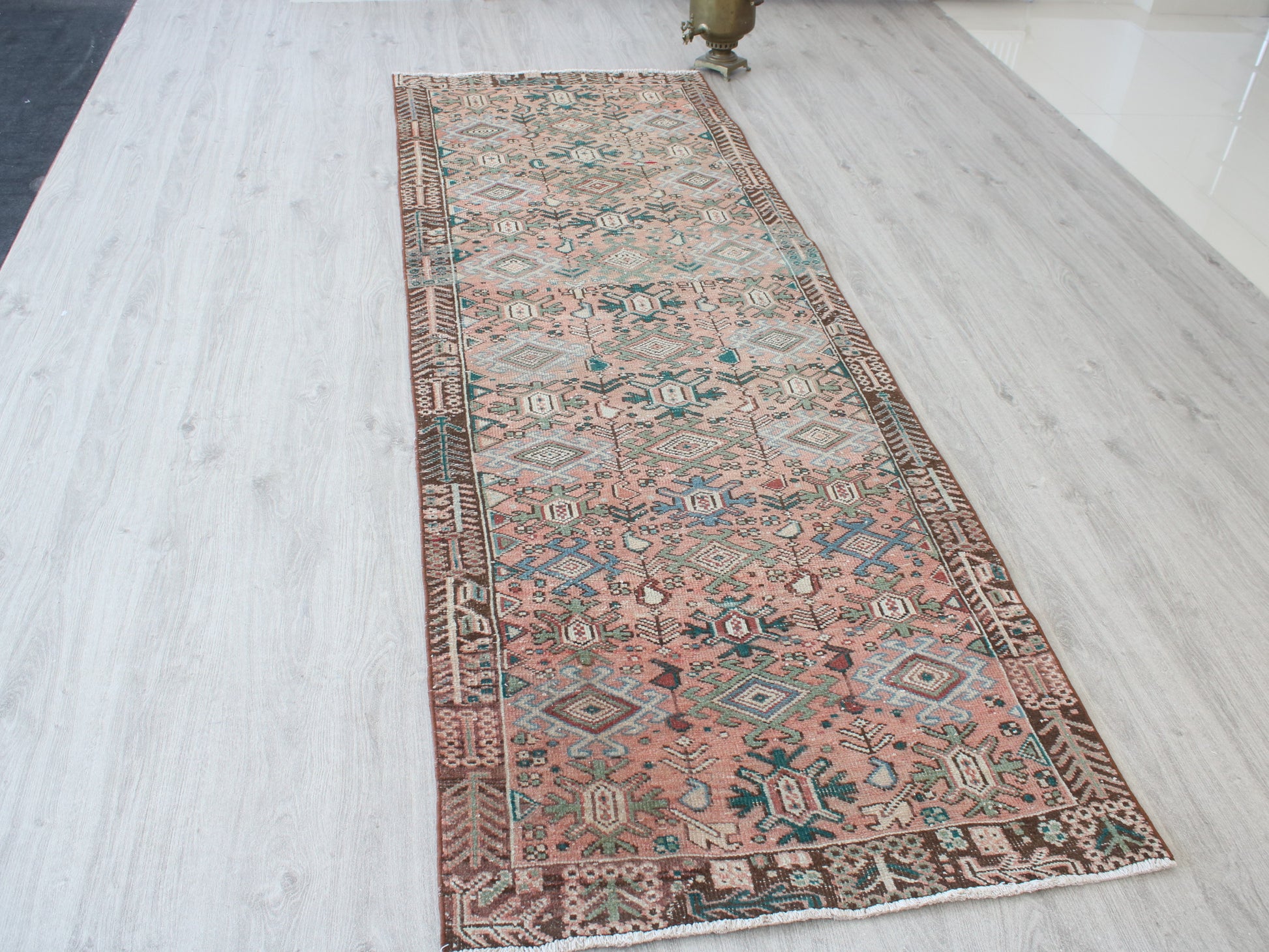 A long runner rug with a traditional Persian-style geometric pattern in dusty pink, brown, teal, and cream, laid out on a light gray wood floor.