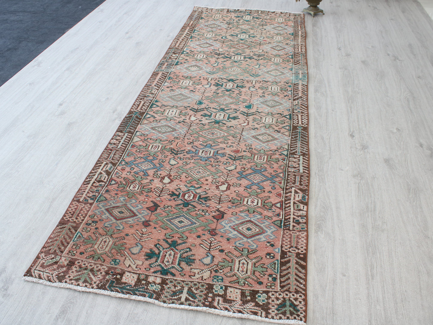 A long rectangular area rug with a distressed geometric pattern in dusty pink, brown, teal, and beige, laid out on a light gray wood floor.