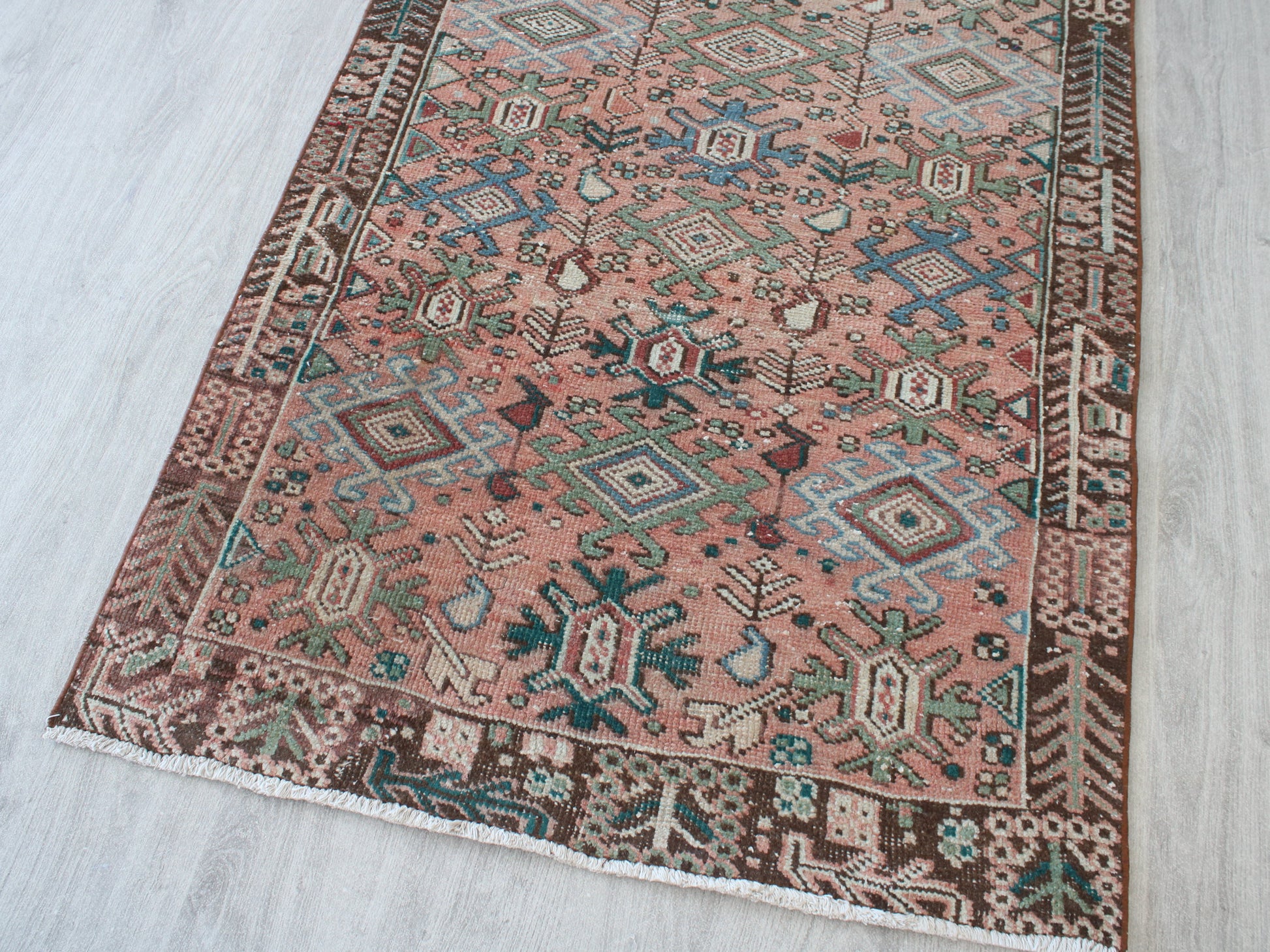 A rectangular area rug with a dusty pink field and intricate geometric borders, laid out on a light gray wood floor.