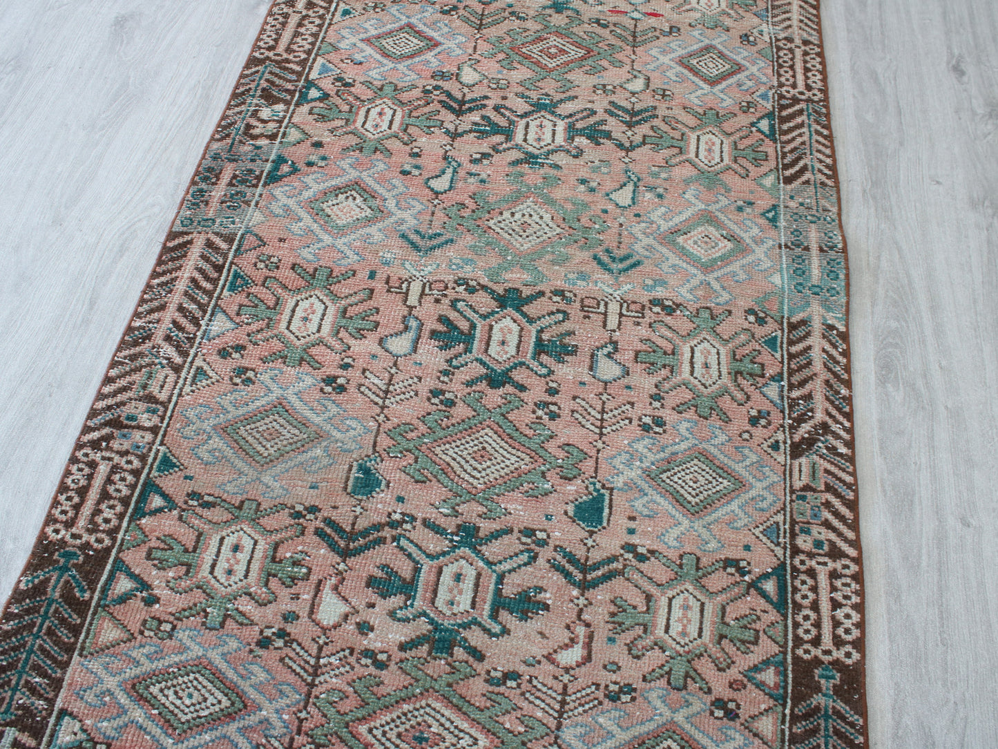 A rectangular area rug with a traditional geometric pattern in dusty pink, teal, brown, and cream, laid out on a light gray wood floor.