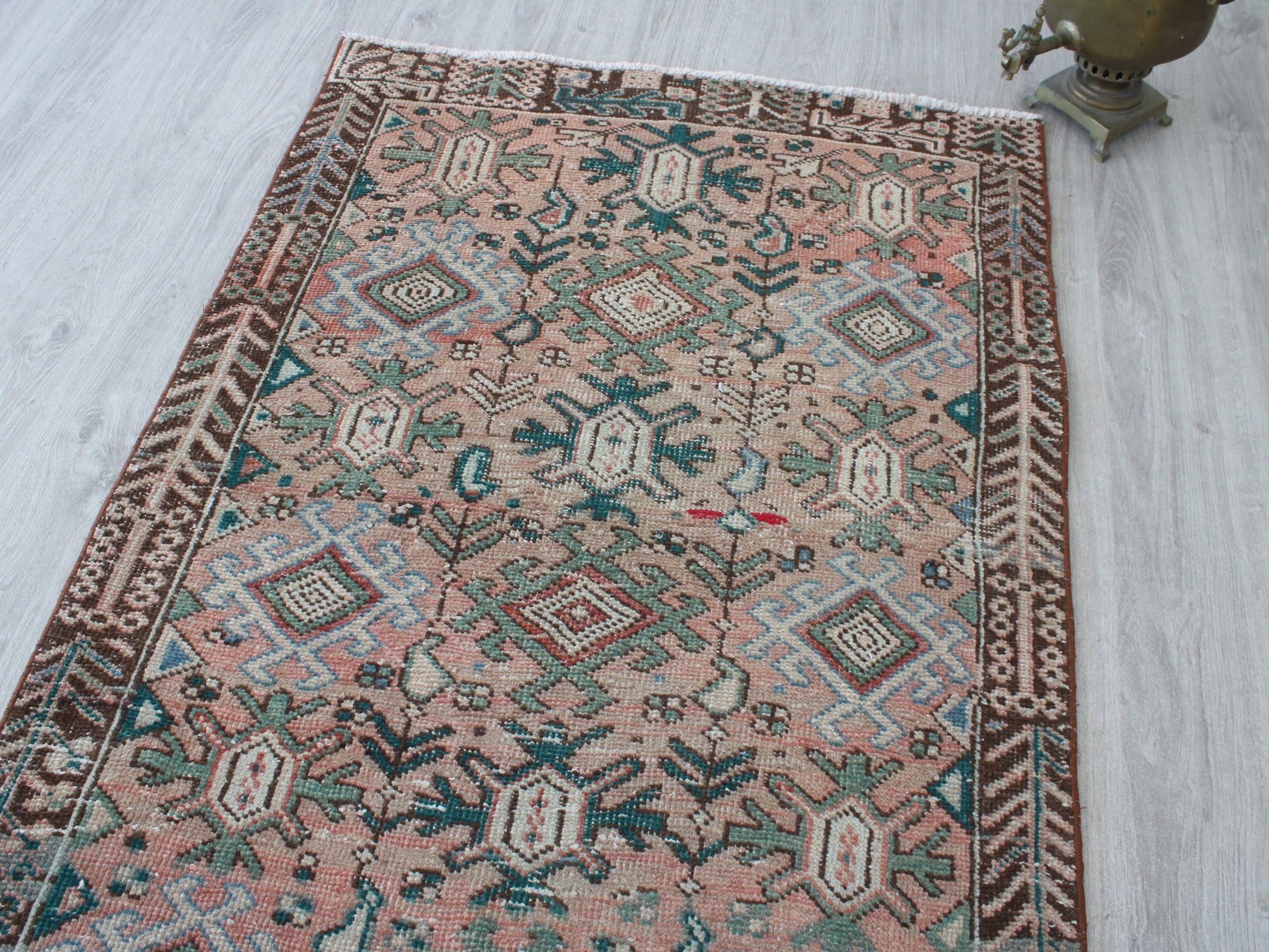 A rectangular vintage-style kilim rug with a dusty pink field, brown borders, and teal and cream geometric motifs, laid out on a light gray wood floor next to a decorative brass object.