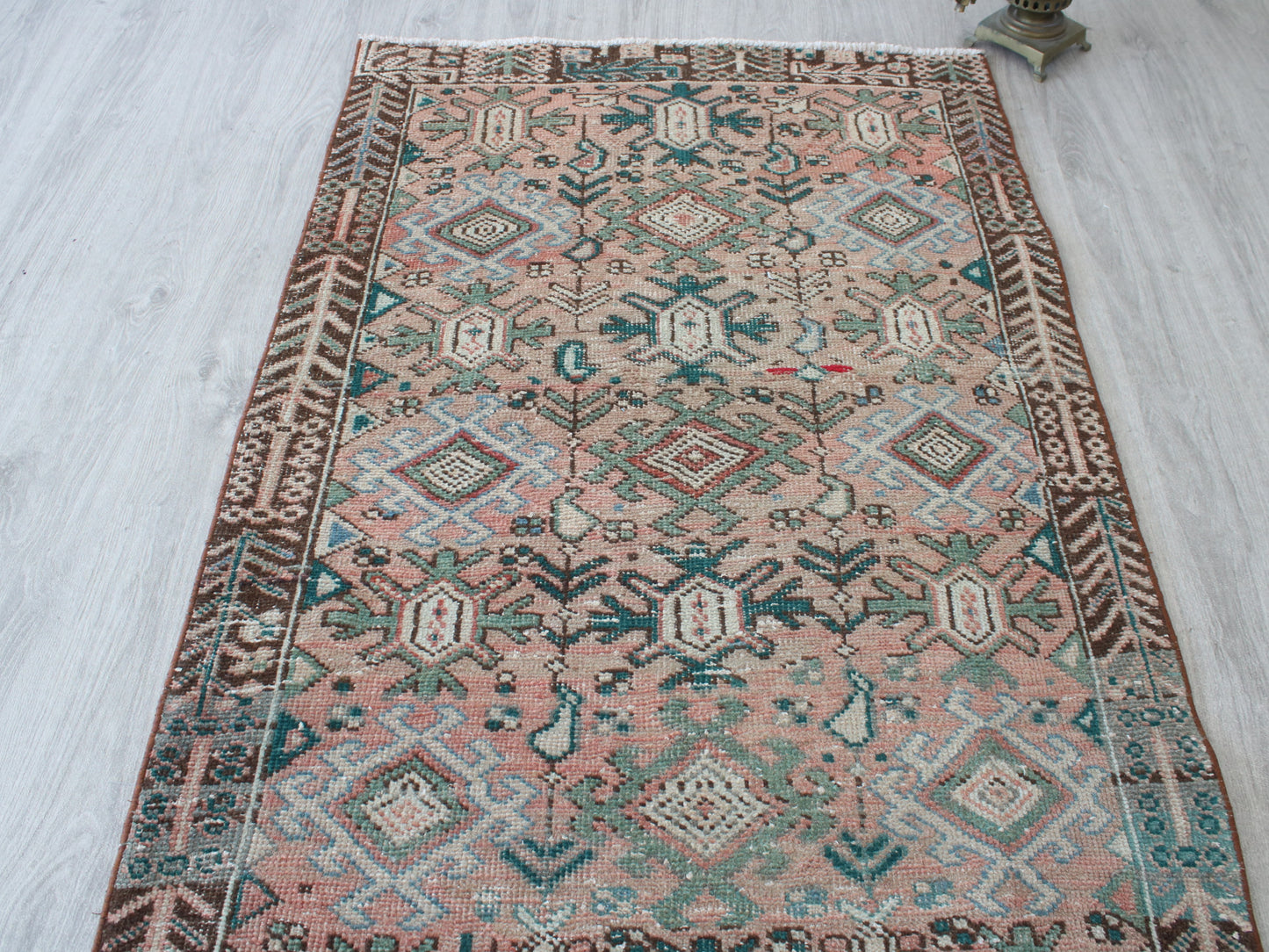 A rectangular vintage-style area rug with a distressed geometric pattern in dusty pink, teal, brown, and cream, laid out on a light gray wood floor.