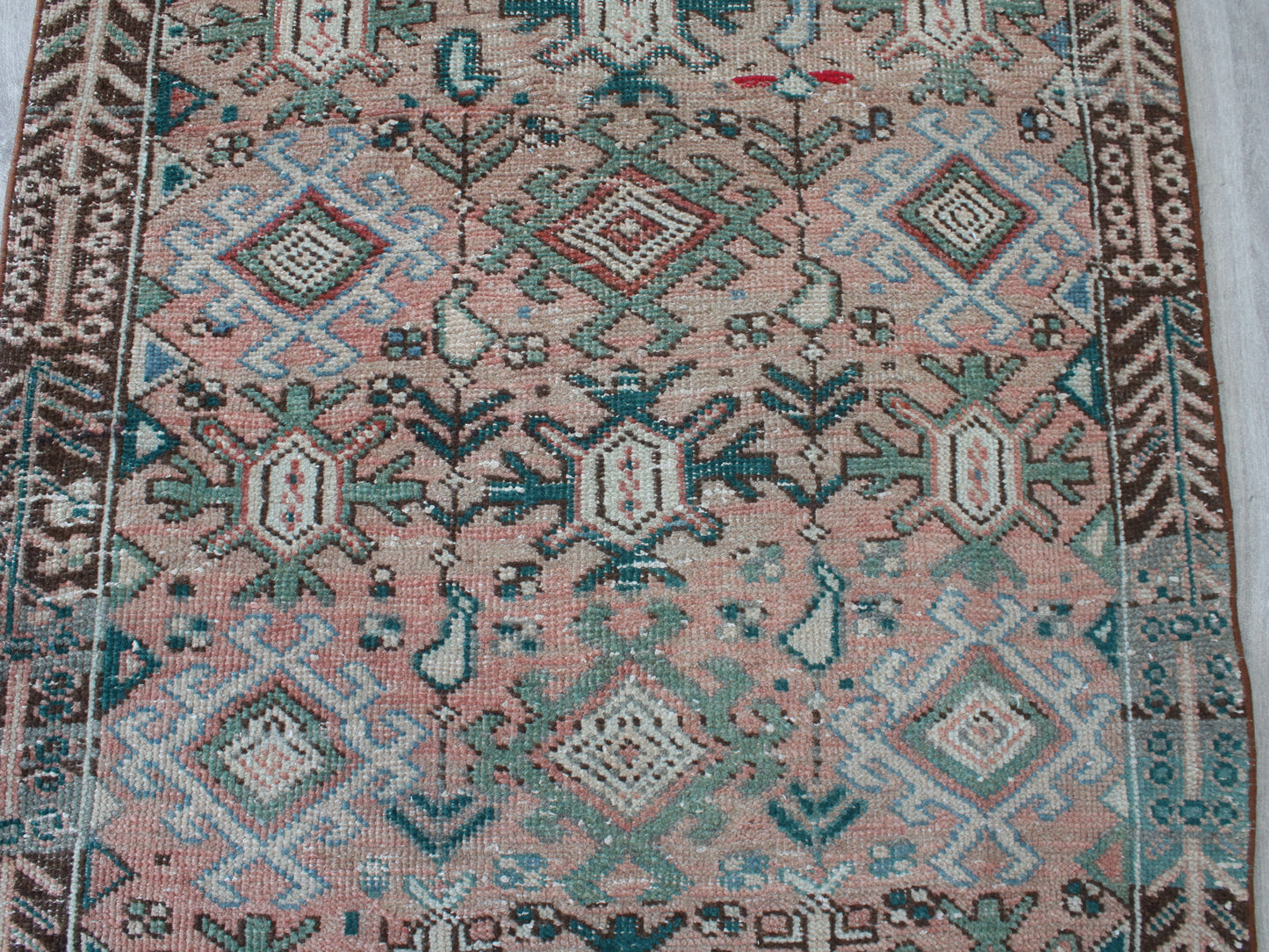 A close-up of a patterned rug with a dusty pink field, featuring geometric and floral motifs in teal, brown, cream, and light blue.