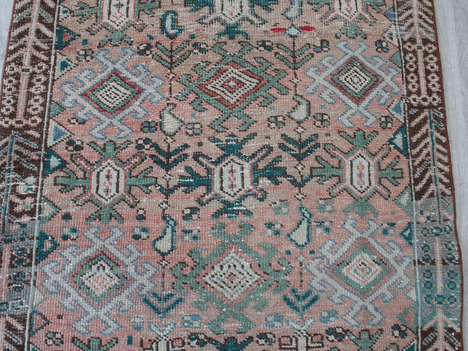 A close-up of a patterned rug with a dusty pink field, featuring geometric and floral motifs in teal, brown, cream, and light blue.