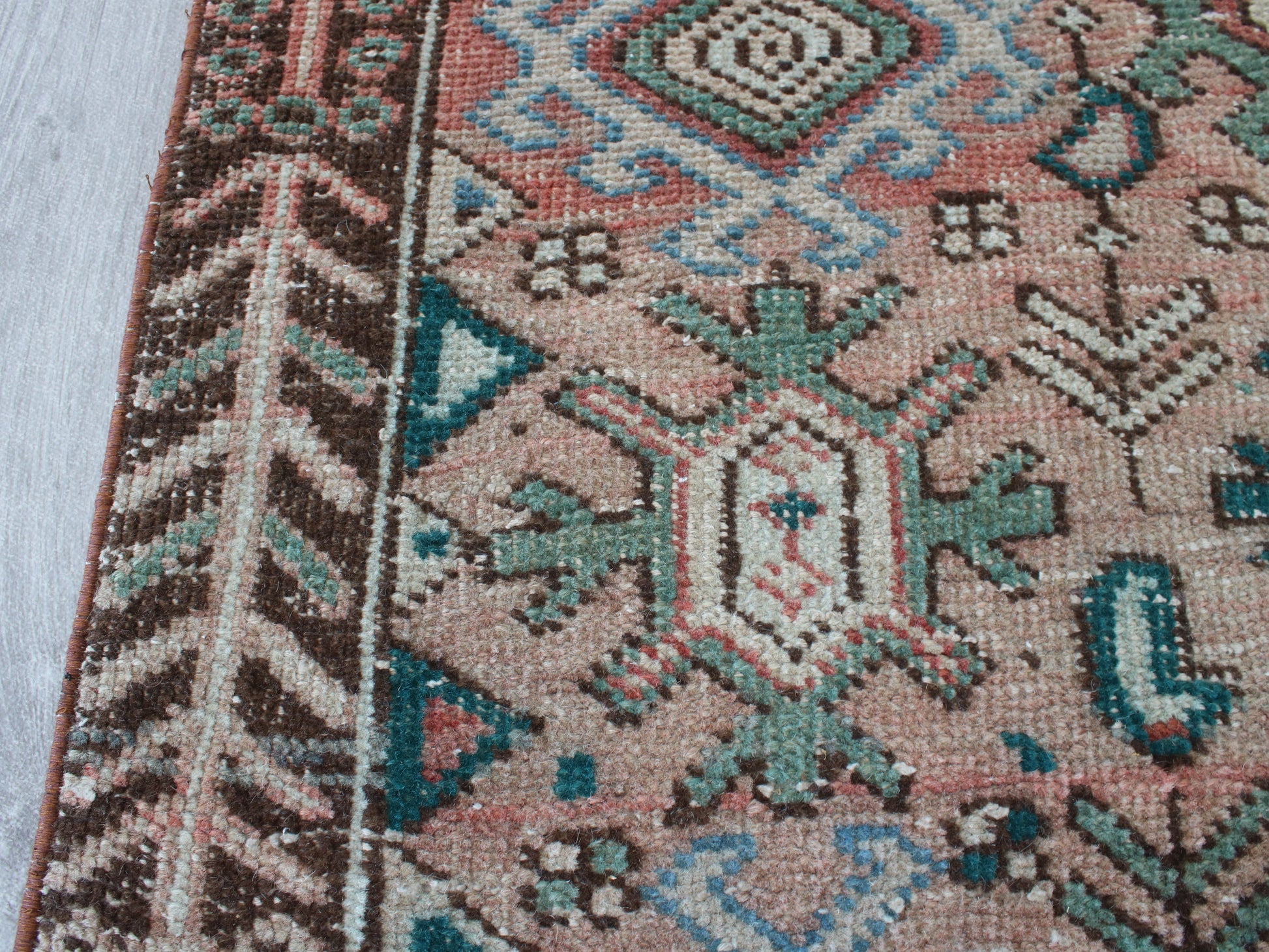 Close-up of a woven rug showing a detailed geometric pattern with a dusty pink field, teal, brown, and cream motifs, and a decorative border.
